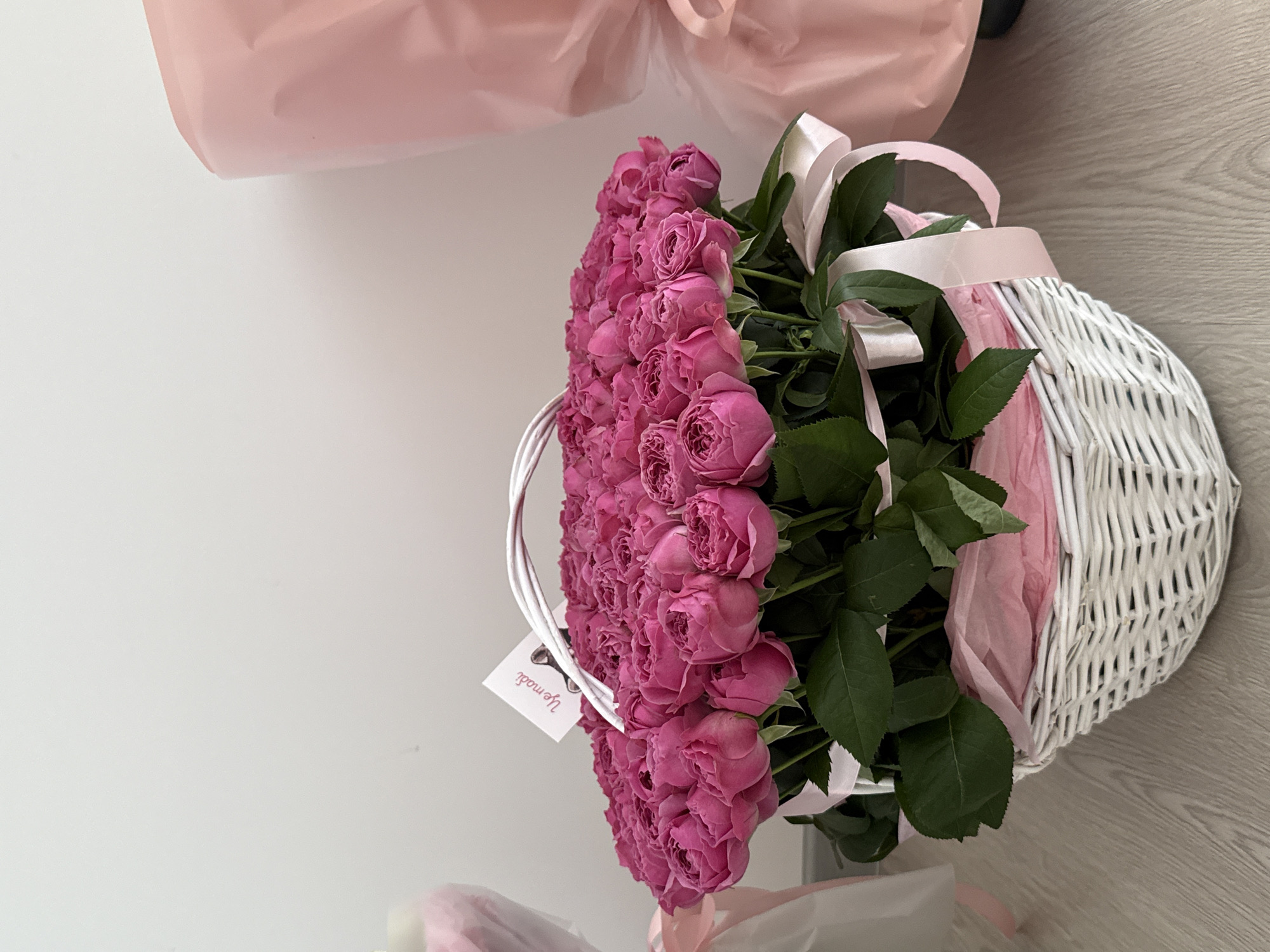 An overhead shot captures a white woven basket brimming with a dense arrangement of bright pink roses. The roses are tightly packed, with their lush green leaves forming a backdrop. A delicate white handle arches over the top of the bouquet, and a small white card with Russian text is tucked amongst the blooms. Soft pink tissue paper lines the inside of the basket, and a pale pink ribbon is tied in a bow on the right side. The basket sits on a light wooden floor, with a pale wall behind it. To the right, partially visible, is a large pink object, possibly gift wrapping. The scene is evenly lit, with no harsh shadows, suggesting diffused lighting conditions. The image appears to be a close-up still life of a floral arrangement.
