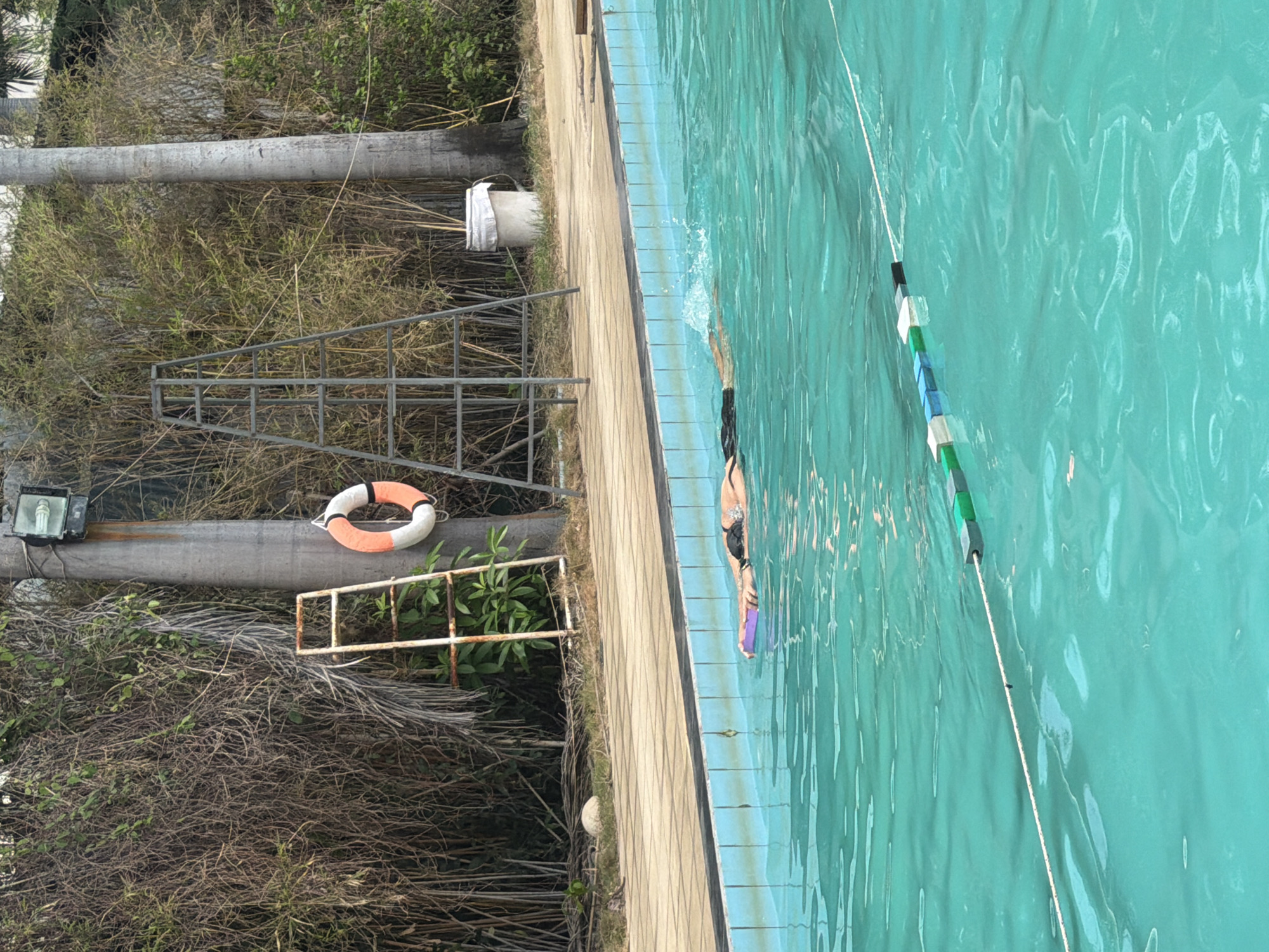 A person swims in a turquoise swimming pool at a resort in Nha Trang, Vietnam. The person is using a purple flotation device and is wearing dark swim trunks. The pool has a lane marker made of buoys. In the background, there is a metal lifeguard stand, a rusted ladder, a life preserver, a trash can, and dense foliage.