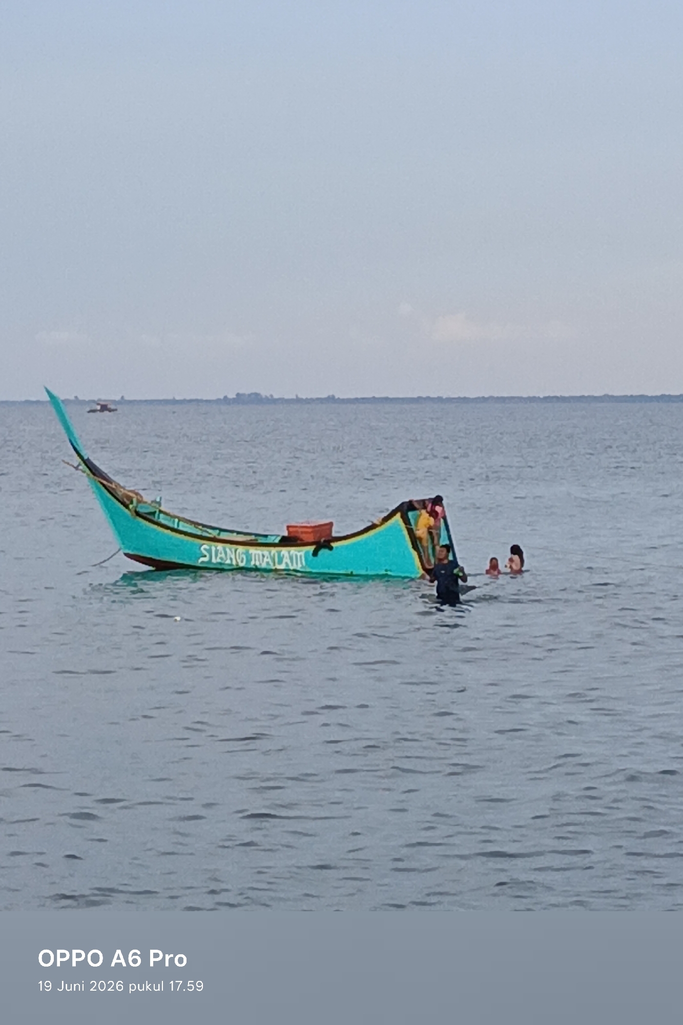 A turquoise-colored longboat is anchored in calm, grey water. The boat is partially submerged, with its bow elevated and stern dipping lower in the water. A man in a dark shirt and shorts stands in the water near the stern, appearing to be assisting or interacting with two people partially visible in the water behind him. One of these individuals is a person with dark hair, and another is a smaller individual whose face is not visible. The boat has the words "SIANG MALAM" painted in white on its side. In the distance, a faint line of land is visible on the horizon, under a hazy sky. A smaller boat is also visible in the far distance. The bottom of the image shows a watermark with "OPPO A6 Pro" and the date and time "19 Juni 2026 pukul 17.59". The scene is likely a coastal area in Lhokseumawe, Indonesia, given the presence of a longboat common in Indonesian waters and the date format.