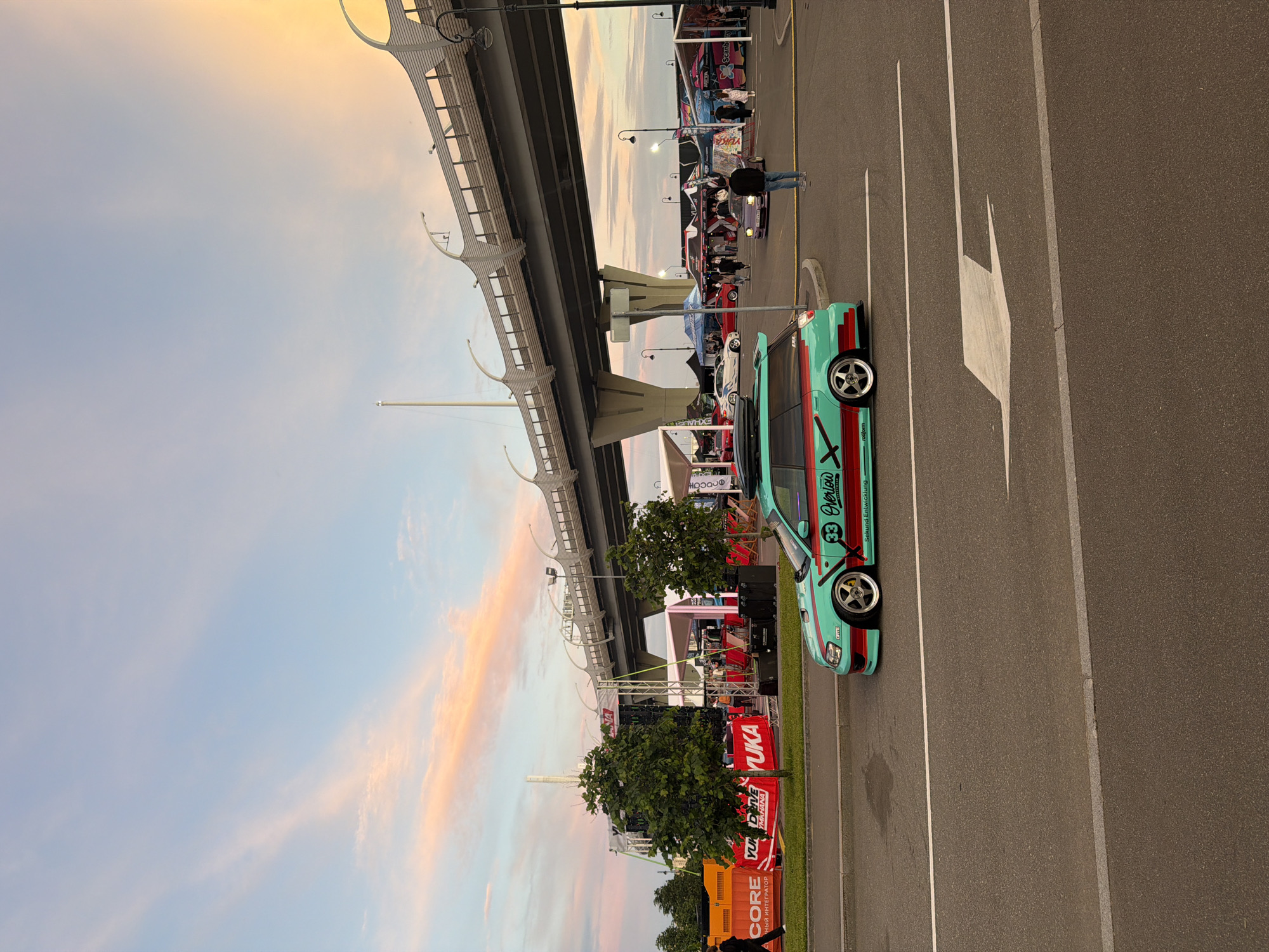 An outdoor scene at dusk shows a turquoise station wagon with red and green racing stripes parked on the side of a road. The car has a roof rack and a custom body kit. In the background, a large elevated highway structure dominates the skyline, with a colorful sunset filling the sky. There are several tents and booths set up under the highway, suggesting a car show or event. A few people are visible walking around the event area. The ground is paved, and a white arrow is painted on the road, indicating a direction. The overall atmosphere is lively and suggests a car enthusiast gathering.