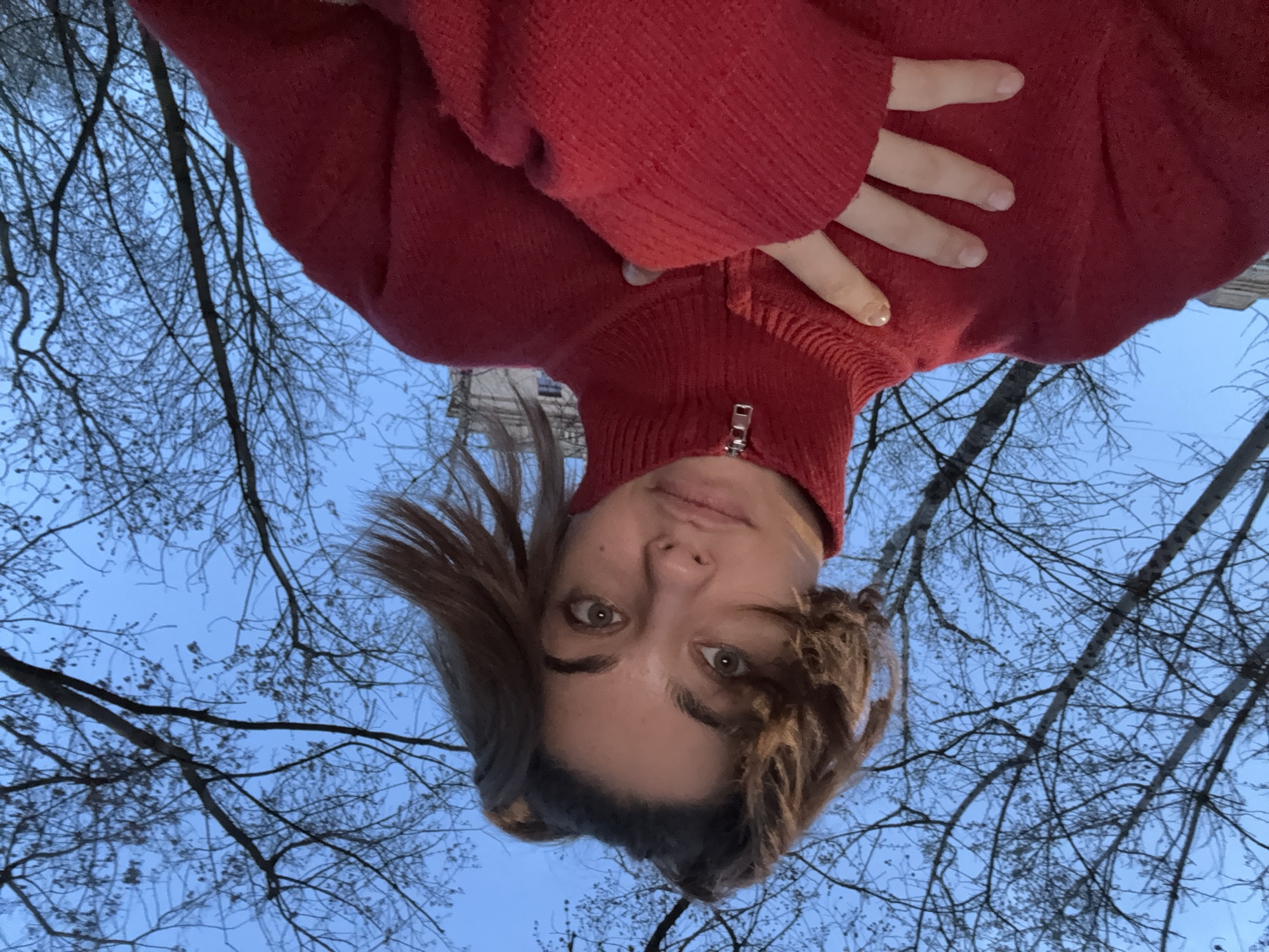 A selfie photograph from a low angle depicts a person with light skin, brown hair, and a red sweater. The person's hand is resting on the sweater's chest area. The background is composed of bare tree branches against a twilight blue sky. The image has been rotated so the subject appears upside down. The exact location within Moscow is not discernible from the visual information provided.