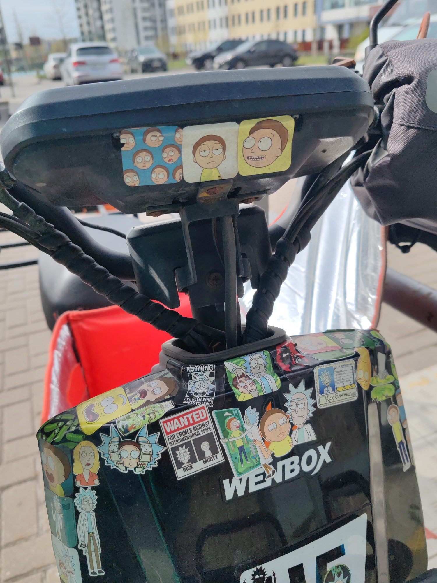 Here's a detailed description of the image:
**Scene:** Close-up of the handlebars and front of a black scooter or electric bike. The scooter is decorated with numerous stickers related to the animated television series "Rick and Morty."
**Details:**
* **Handlebars:** The handlebars are covered with black wrapped cables.
* **Stickers:** The scooter is heavily decorated with "Rick and Morty" themed stickers, including characters such as Rick, Morty, Summer, and various scenes from the show. One sticker reads "WANTED FOR CRIMES AGAINST INTERDIMENSIONAL SPACE", and another reads "WENBOX". Another reads "NOTHING! I'VE SEEN WHAT MAKES YOU VEER" and features Rick Sanchez. Another shows Rick's driver's license.
* **Objects:** There is a silver reflector visible. Part of a red delivery box is visible in the background.
**Environment:**
* **Location:** The scooter is parked on a paved area.
* **Background:** Buildings are visible in the blurred background. Cars are visible in the distance.
* **City:** The picture was most likely taken in Saint Petersburg, Russia.