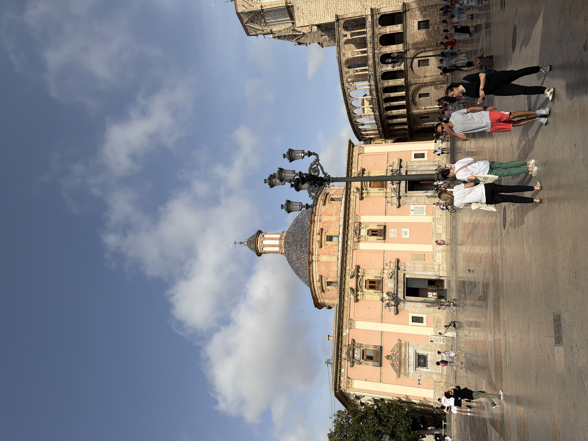 The image shows a wide shot of a public square in front of a large pink building with a dome and an adjoining stone structure with arches. The sky is blue with scattered white clouds. Several people are walking and standing in the square. In the foreground, a group of four people are standing together. A man in a grey t-shirt and red shorts is facing right, with his hand near his mouth. A woman in a black t-shirt and black pants is facing left. Two women in white tops and black pants are also in the foreground, facing right. A black lamppost with multiple lights stands in the middle of the square. Other people are visible in the background, as well as trees and architectural elements of the buildings. The ground of the square appears to be paved with stone tiles.