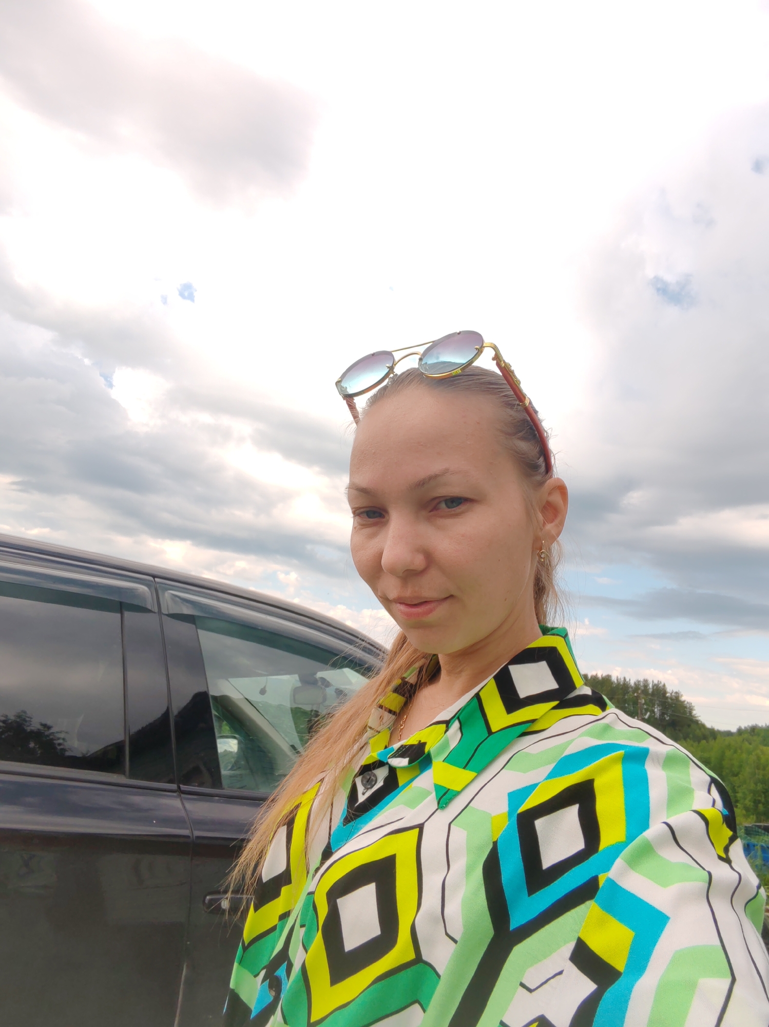A medium shot portrait shows a young woman standing next to a black car under a cloudy sky. The woman has blonde hair and is wearing sunglasses perched on her head. She is dressed in a brightly colored shirt with a geometric pattern of white, black, green, and yellow. The car is dark and appears to be a four-door sedan. The background suggests an outdoor setting, possibly a roadside or parking area, with trees visible in the distance. The overall impression is a casual, outdoor snapshot. No specific details about the location in Чухлэм, Russia, can be determined from the image.