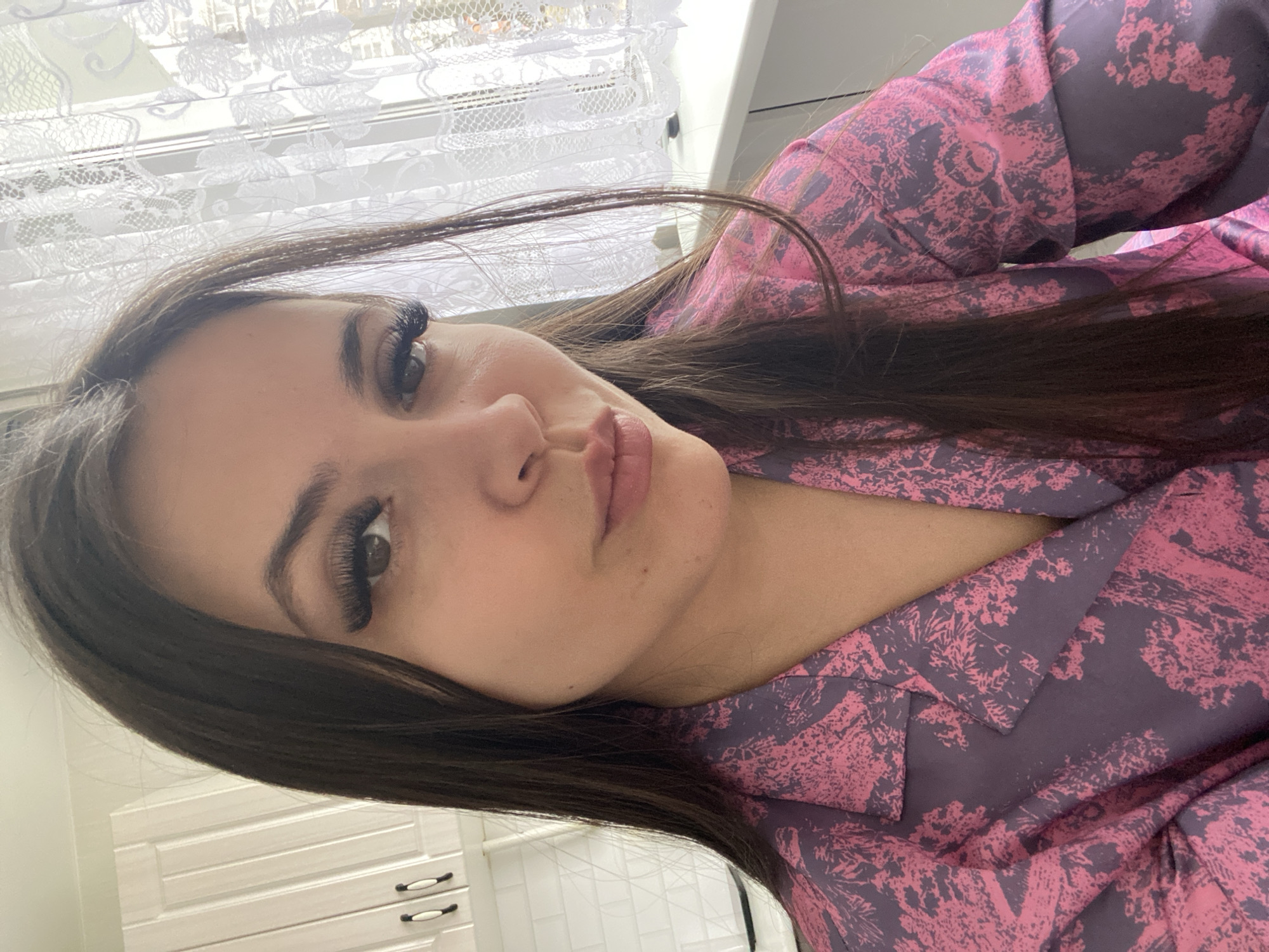 A young woman with long, dark brown hair, is indoors with a white window and white cabinets behind her. She is wearing a pink and purple patterned shirt. Her eyes are dark and she has long eyelashes.