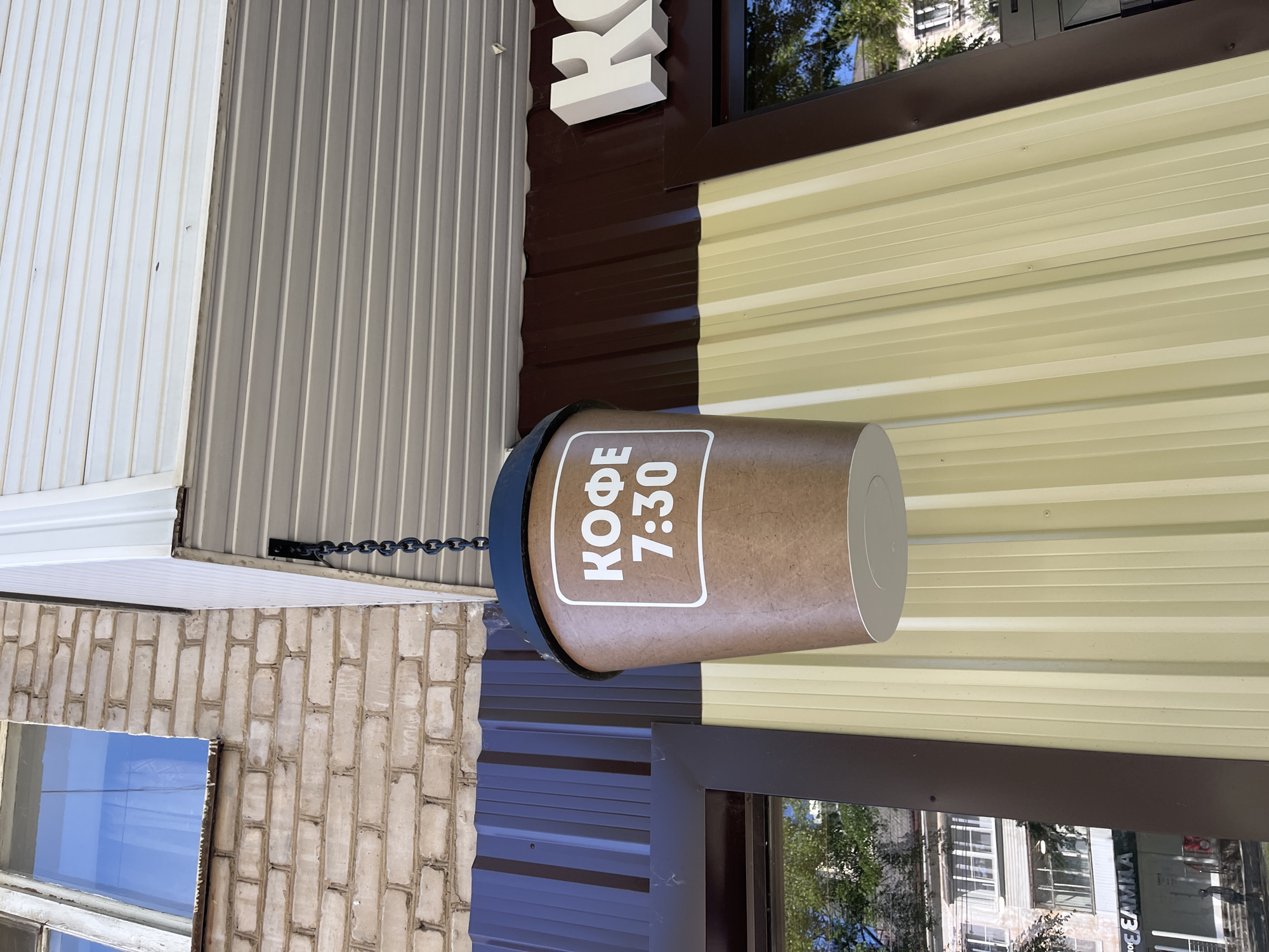 A close-up, eye-level, outdoor shot captures a sign for "COFFEE 7:30" in Chaikovsky, Russia. The sign is shaped like a large paper coffee cup and is hanging from a chain under a white, paneled overhang. The cup is brown with white text within a white outlined box. Behind the cup, a building wall made of light-colored brick is visible. To the right of the cup, a dark brown, corrugated metal wall is present, and further to the right, large white 3D letters spelling "K" are mounted. A light yellow, corrugated metal wall is also visible below the overhang. The scene is illuminated by daylight, suggesting it is daytime.