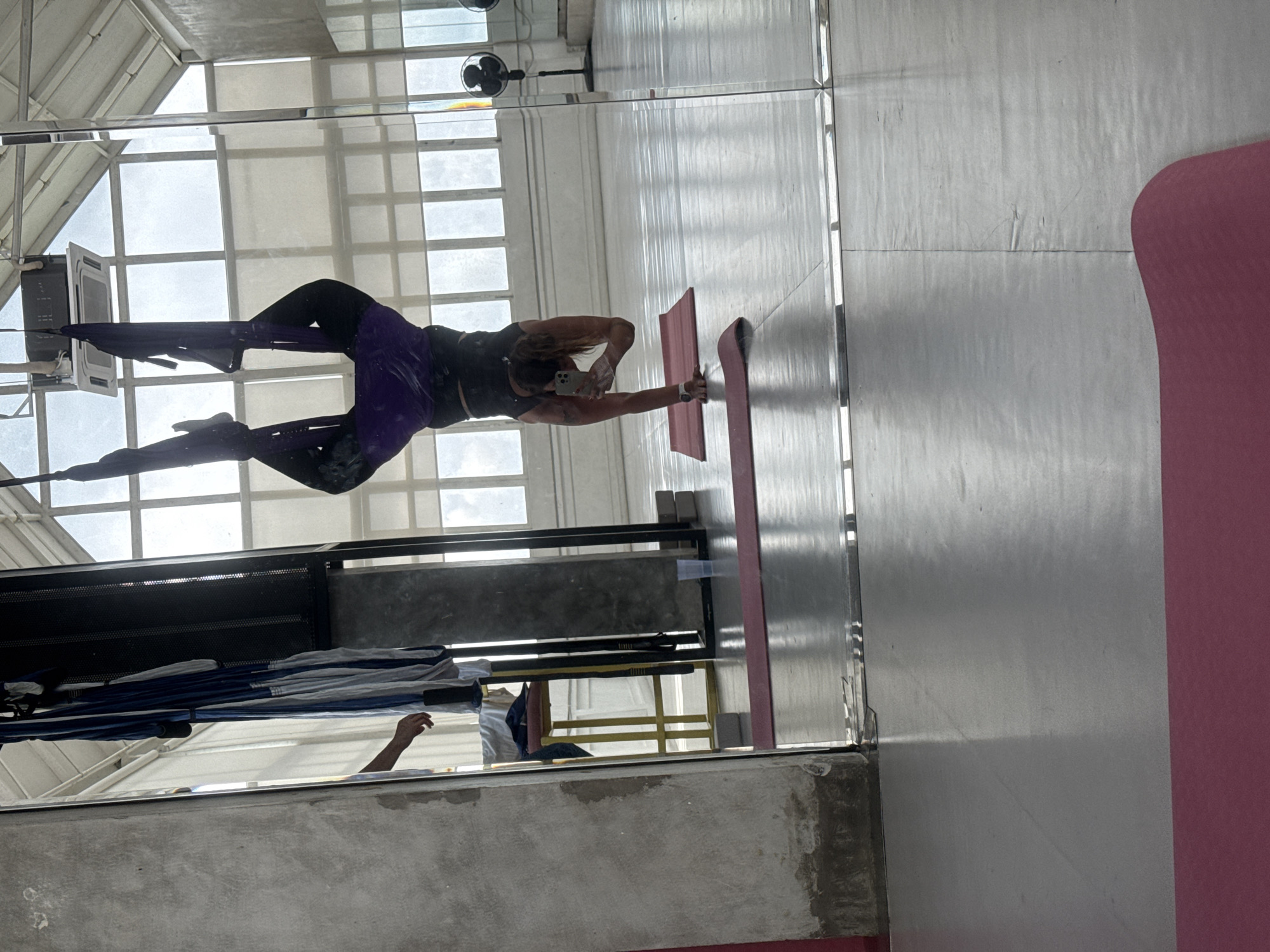 The image shows a woman in a mirrored studio doing aerial yoga, location Sukawati, Indonesia. The woman is wearing dark leggings and a dark top, and is suspended from fabric attached to the ceiling. She is holding her phone, possibly taking a picture of herself in the mirror. There are yoga mats on the floor and a fan mounted on the ceiling near the windows. The studio has large windows that let in natural light. There is a metal structure on the ceiling, perhaps used for aerial equipment.