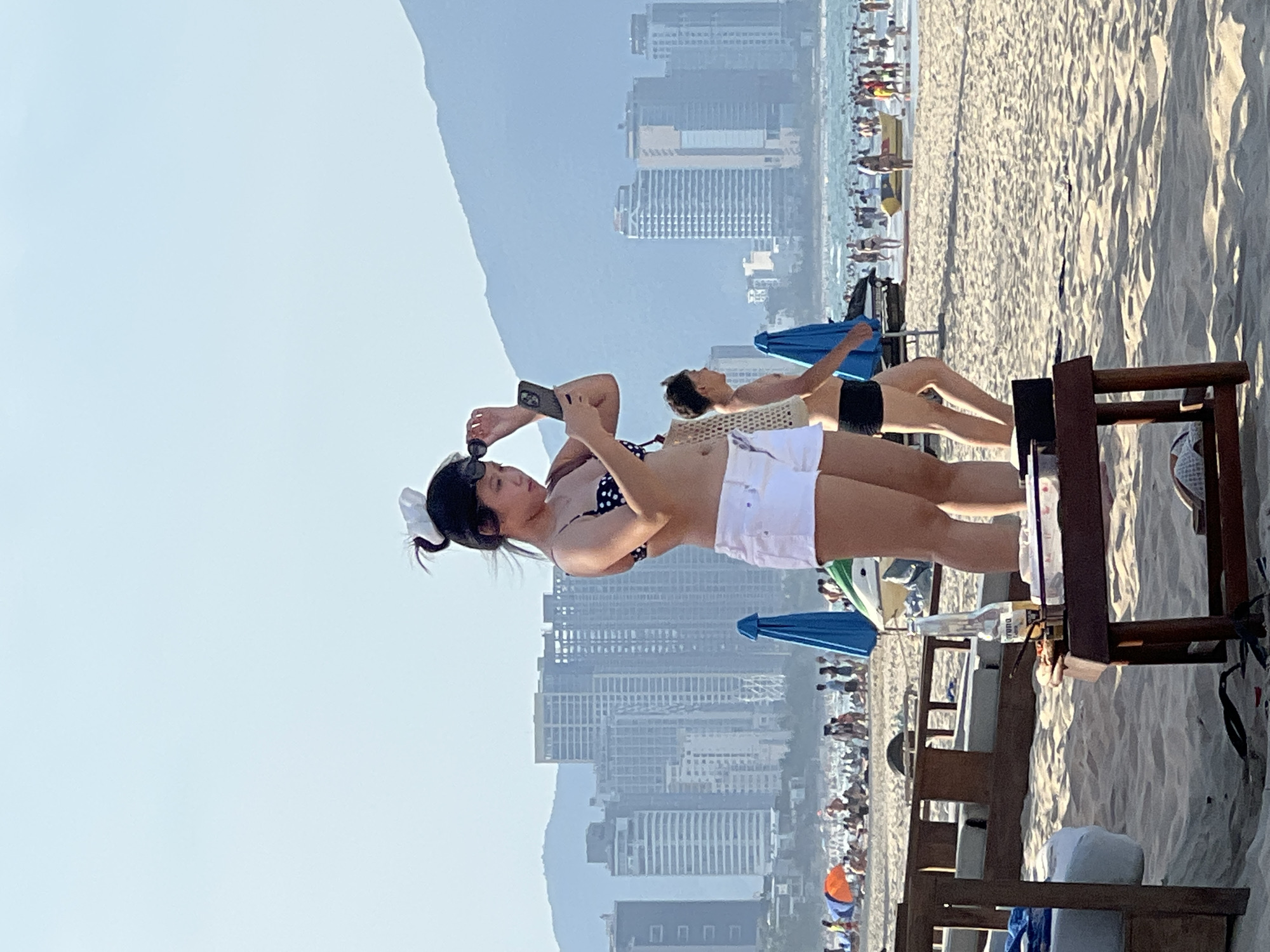 The image depicts a beach scene in Da Nang, Vietnam, with a focus on a young woman in the foreground. She is wearing a polka dot bikini top and white shorts, holding sunglasses in her right hand and a smartphone in her left, appearing to adjust her sunglasses or check her phone. Her hair is pulled up in a bun with a white accessory.
Behind her, a partially visible young man in black swim trunks is walking away from the camera. In the middle ground, there are beach chairs, umbrellas, and a table with a bottle of beer and some food items. The background reveals a bustling beach with many people swimming, sunbathing, and engaging in recreational activities.
Further in the distance, the city skyline of Da Nang is visible, characterized by numerous high-rise buildings and mountains. The sky is clear and bright, suggesting a sunny day. The overall scene conveys a lively atmosphere of a popular beach resort.