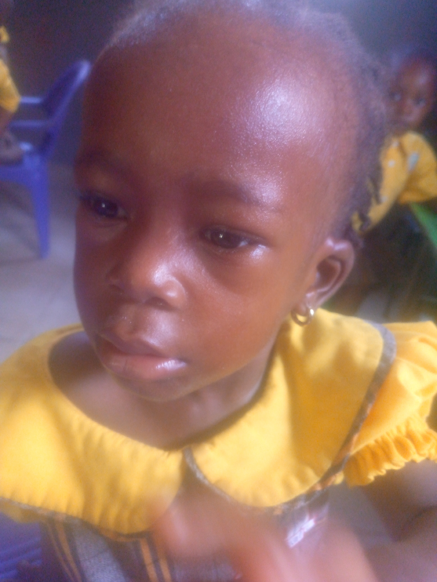 A close-up shot of a young, dark-skinned child with an earring, wearing a yellow shirt with a ruffled collar. Another child, also in yellow, is visible in the background, partially obscured. A blue plastic chair is also visible. The location, Ikorodu, Nigeria, is mentioned as a city only, it is not evident within the image. No specific actions or interactions can be determined from the image. The environment appears to be indoors.