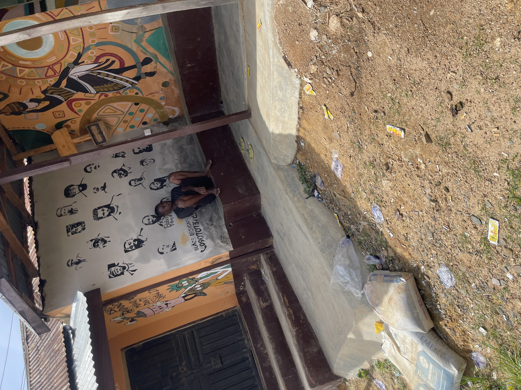 The image depicts an outdoor scene in Jos, Nigeria, with a building exhibiting a vibrant mural. On the left side of the building, an orange-colored section features cartoonish figures, possibly from a popular animated series, painted in black and green. Adjacent to this, a white wall is adorned with numerous black-and-white stencil portraits of people. A young Black male, wearing a dark t-shirt and shorts, is seated on a low concrete ledge in front of the white wall. His legs are crossed, and he is looking towards the right of the frame. Further to the right, the building displays a large, colorful mural with an African-inspired design, featuring a central figure carrying a pot on her head. The ground in front of the building is a mix of dirt and concrete. Scattered on the ground are various items, including two pairs of yellow flip-flops, several small pieces of litter such as wrappers and plastic bags, and two large sacks of cement. The overall environment appears somewhat unkempt, with grass growing in patches around the dirt and rubble. The building has a corrugated metal roof, supported by wooden beams.