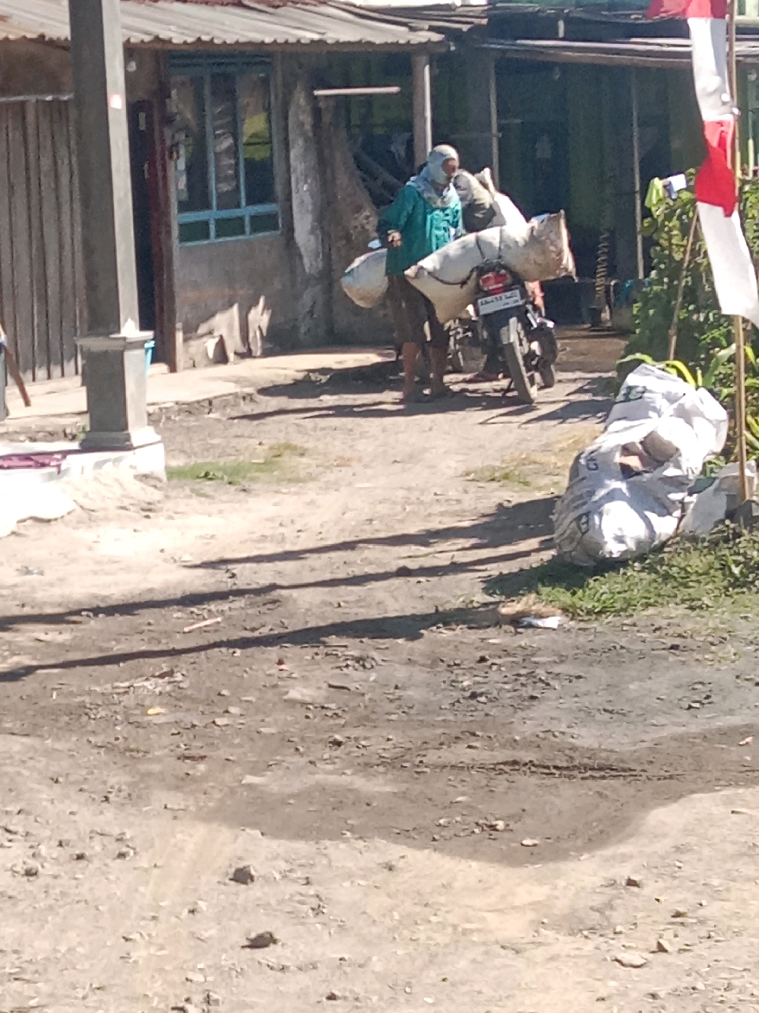 In the city of Jurangjeru, Indonesia, a scene unfolds on a dusty, uneven ground. A person, possibly a woman, is dressed in a turquoise jacket and head covering, standing next to a motorcycle. The motorcycle is laden with two large, white sacks, secured on the rear seat and possibly a rack. One sack appears to be bulging, while the other is more elongated. In the background, a weathered building with a corrugated metal roof is visible, featuring a door and a window with blue frames. Adjacent to the building, the foliage is green and somewhat overgrown. To the right, a tall, thin structure, possibly a flagpole, displays a partially unfurled Indonesian flag with its characteristic red and white bands. Another large white sack rests on the ground near the flag. The overall environment suggests a rural or semi-urban setting.