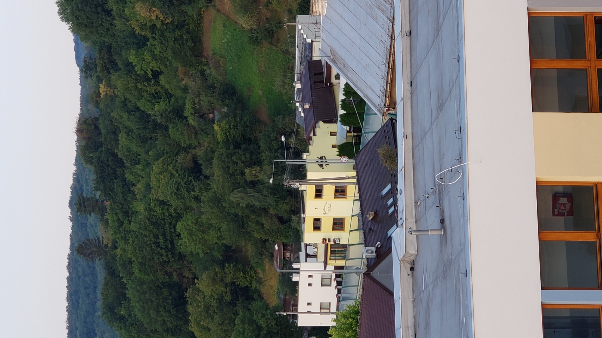 The image captures an outdoor scene with a residential and commercial area in the foreground and a densely wooded hillside in the background. A light-colored building with multiple windows is visible in the middle ground, painted yellow, with signage indicating "Reštaurácia Amadea". Adjacent to it is a white building with several windows. Several roofs are visible in the foreground, some with a dark, sloped design and others with a lighter, flat material. A white cable is coiled on the flat roof in the immediate foreground, attached to a metal post. The background is dominated by a lush green forest that covers a rolling hill. The sky is clear and pale, suggesting it might be early morning or late afternoon. The scene appears to be a section of the city of Prešov, Slovakia, with its characteristic mix of urban structures nestled against natural landscapes.
