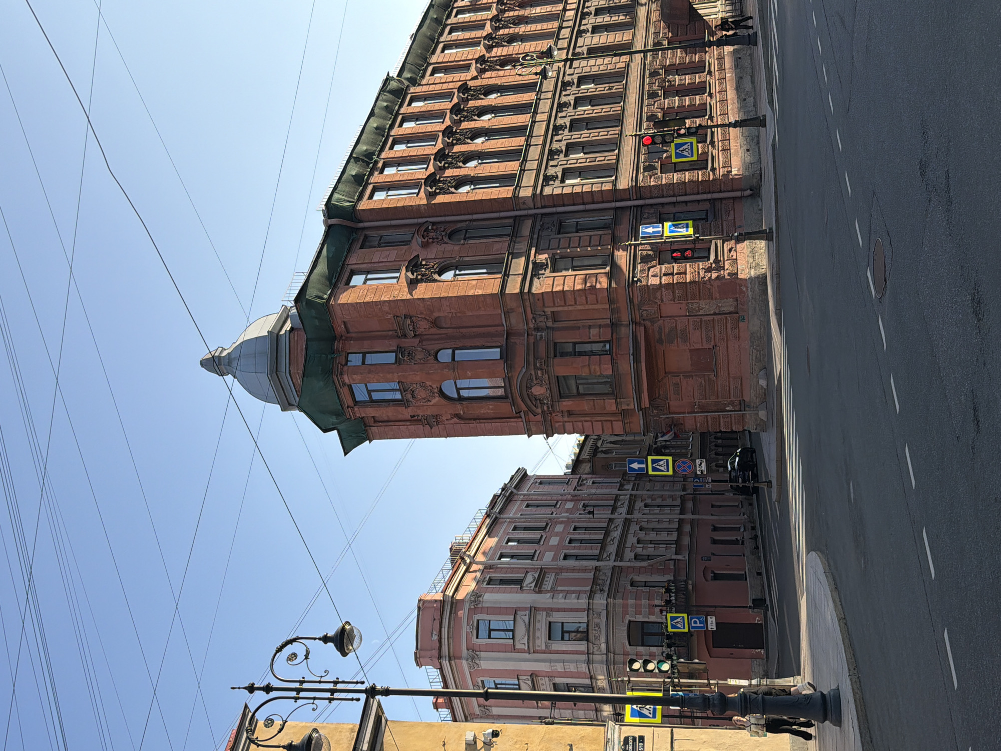 This is a low-angle, medium shot captured in Saint Petersburg, Russia, on a clear, bright day. The scene depicts a street intersection with ornate, historic brick and stucco buildings on either side.
In the foreground, the asphalt street features white dashed lines and a circular manhole cover. A pedestrian crossing with white stripes is visible on the right. The sidewalk on the left is paved with light gray concrete.
Two women are walking on the sidewalk on the left. One woman, wearing a light-colored jacket and dark pants, is closer to the foreground, and another woman in dark clothing is behind her.
Several streetlights with decorative black metal poles are present. One prominent streetlight on the left is partially obscuring a yellow sign with a pedestrian symbol and a "P" indicating parking.
Traffic signals are visible, including a set of green and red lights on the left and a red light on the right. Several yellow pedestrian crossing signs with blue directional arrows are also positioned at the intersection.
The buildings are characterized by their detailed architectural features. The building on the right is a tall, red brick structure with a domed roof section and numerous windows with decorative molding. The building on the left is a pink stucco building with white trim and many windows.
Overhead, numerous power lines crisscross the clear blue sky. A black car is parked or driving on the street in the background between the buildings. The lighting suggests it is either early morning or late afternoon, with long shadows cast by the buildings and streetlights. The overall impression is one of urban architecture and city life in a historic setting.