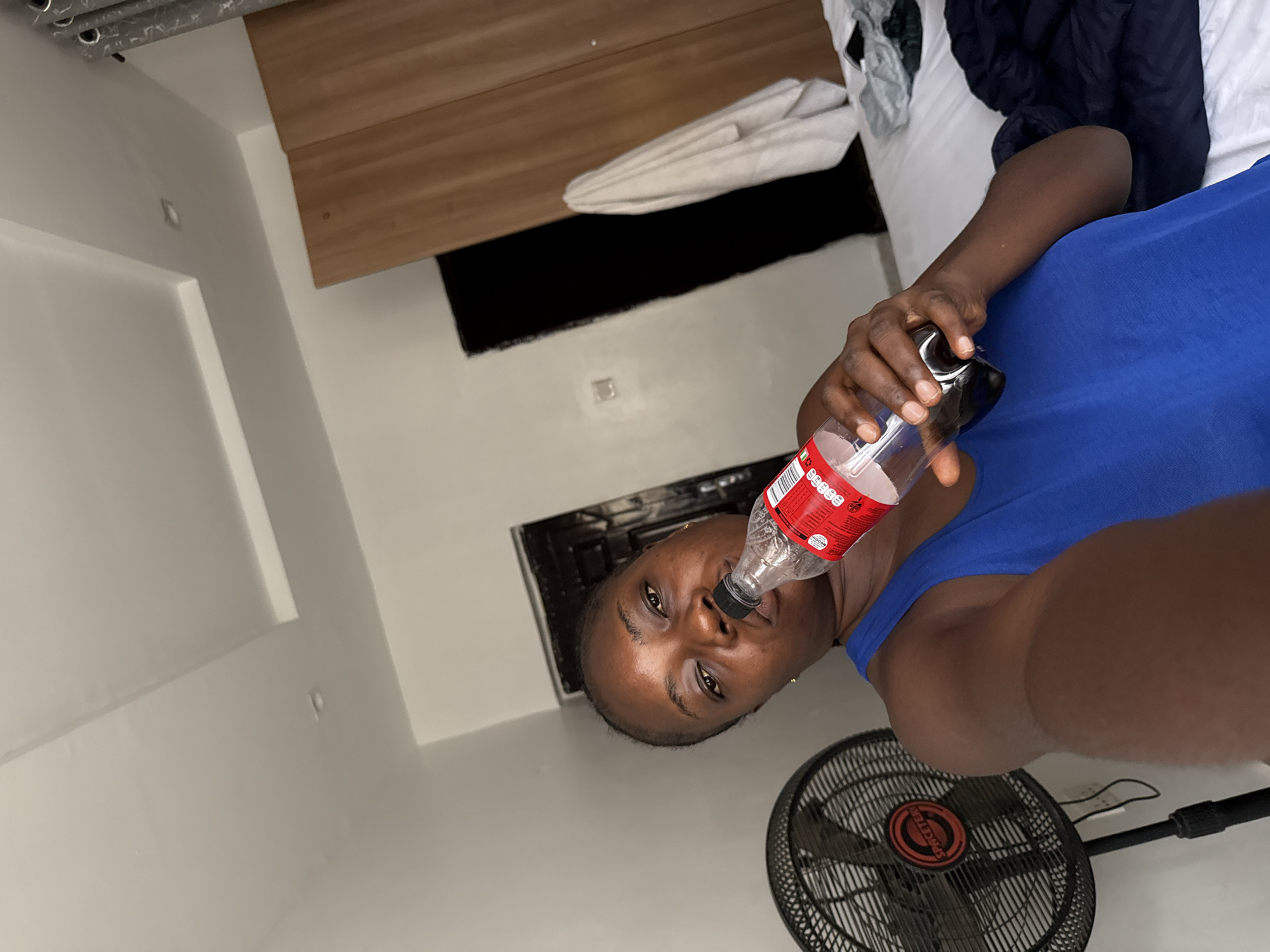 A person is holding a plastic bottle to their mouth, presumably drinking from it. The person is wearing a blue shirt. A black fan is visible in the lower right of the frame. The location appears to be indoors, likely a bedroom, as there is a bed with bedding visible in the background. There is a wooden paneling feature on the wall. The exact location in the city of Iddo Tudunwada, Nigeria, cannot be determined from the image.