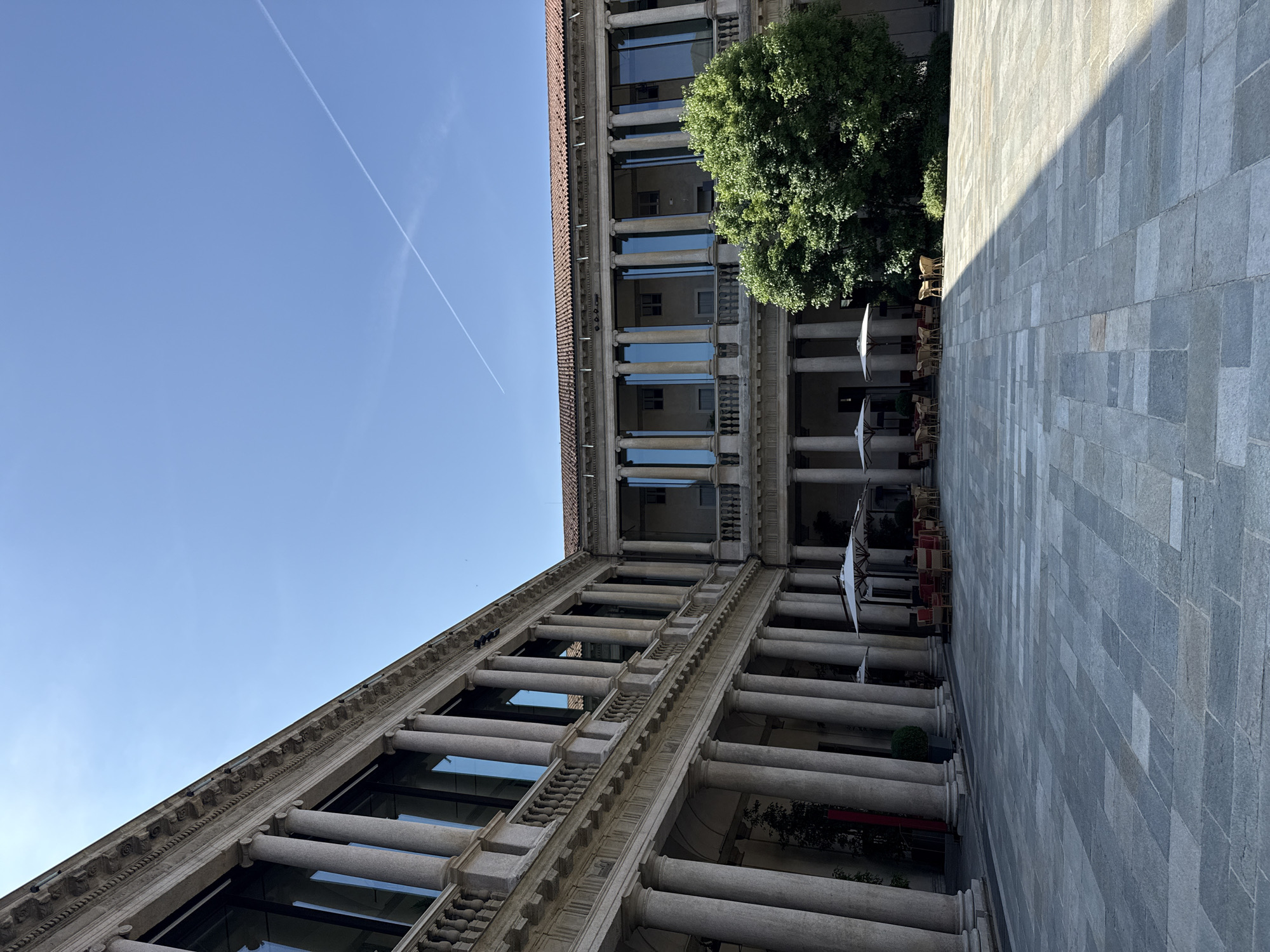 The image captures a courtyard in Milan, Italy, showcasing classical architectural elements. The perspective is from below, looking upwards at a building facade composed of multiple stories with rows of columns and arched windows. The sky is visible in the upper left, a clear blue with a contrail. To the right, a mature tree partially obscures the building. Below, there is an open-air space, likely a restaurant or café, with tables and umbrellas. The ground is paved with grey rectangular stones. The image does not contain people.