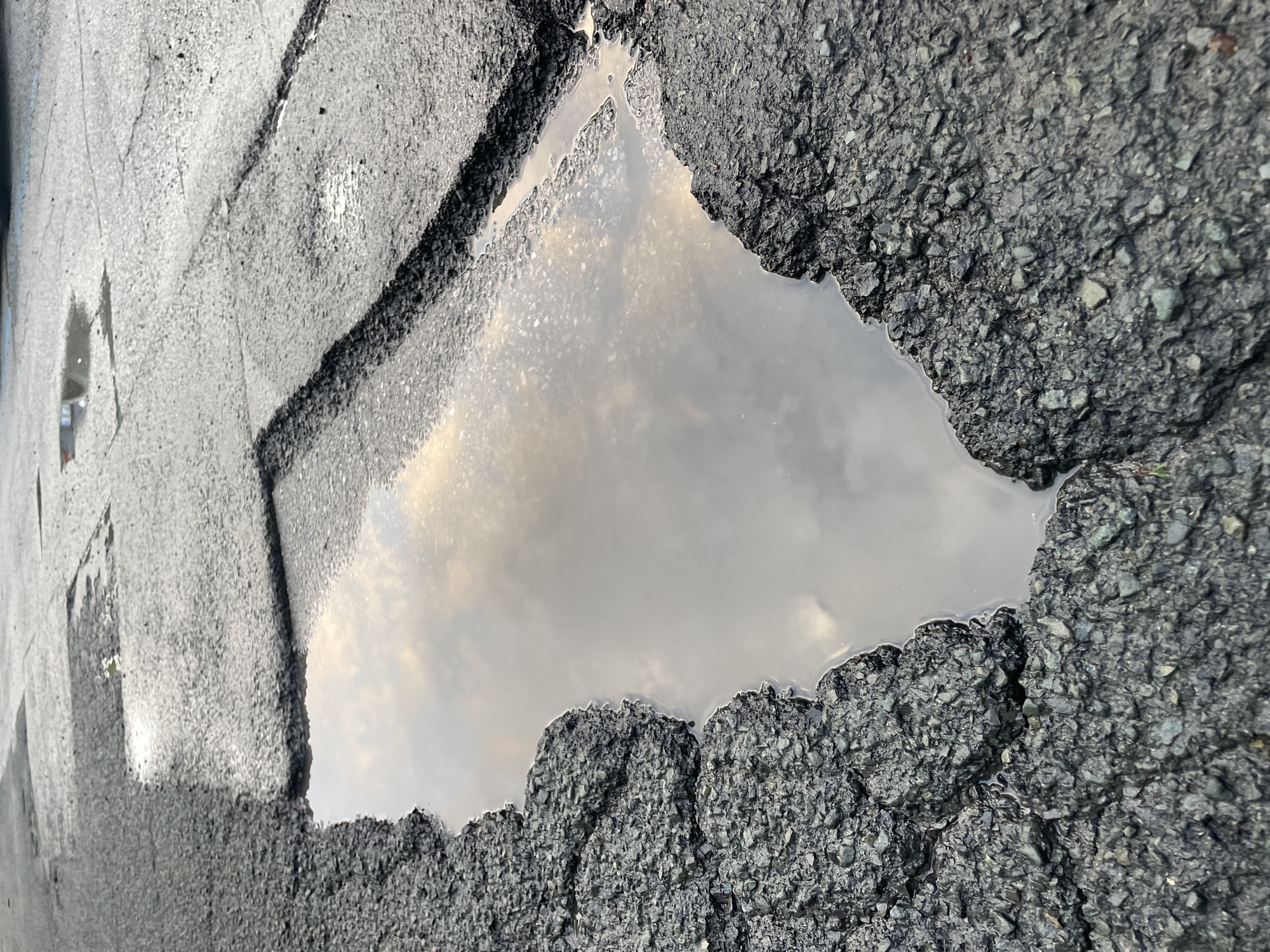 A close-up, angled view of a water-filled pothole in asphalt pavement. The asphalt surrounding the pothole shows signs of wear and cracking. The water surface reflects a cloudy sky. The pothole is located in a street in Burgas, Bulgaria.