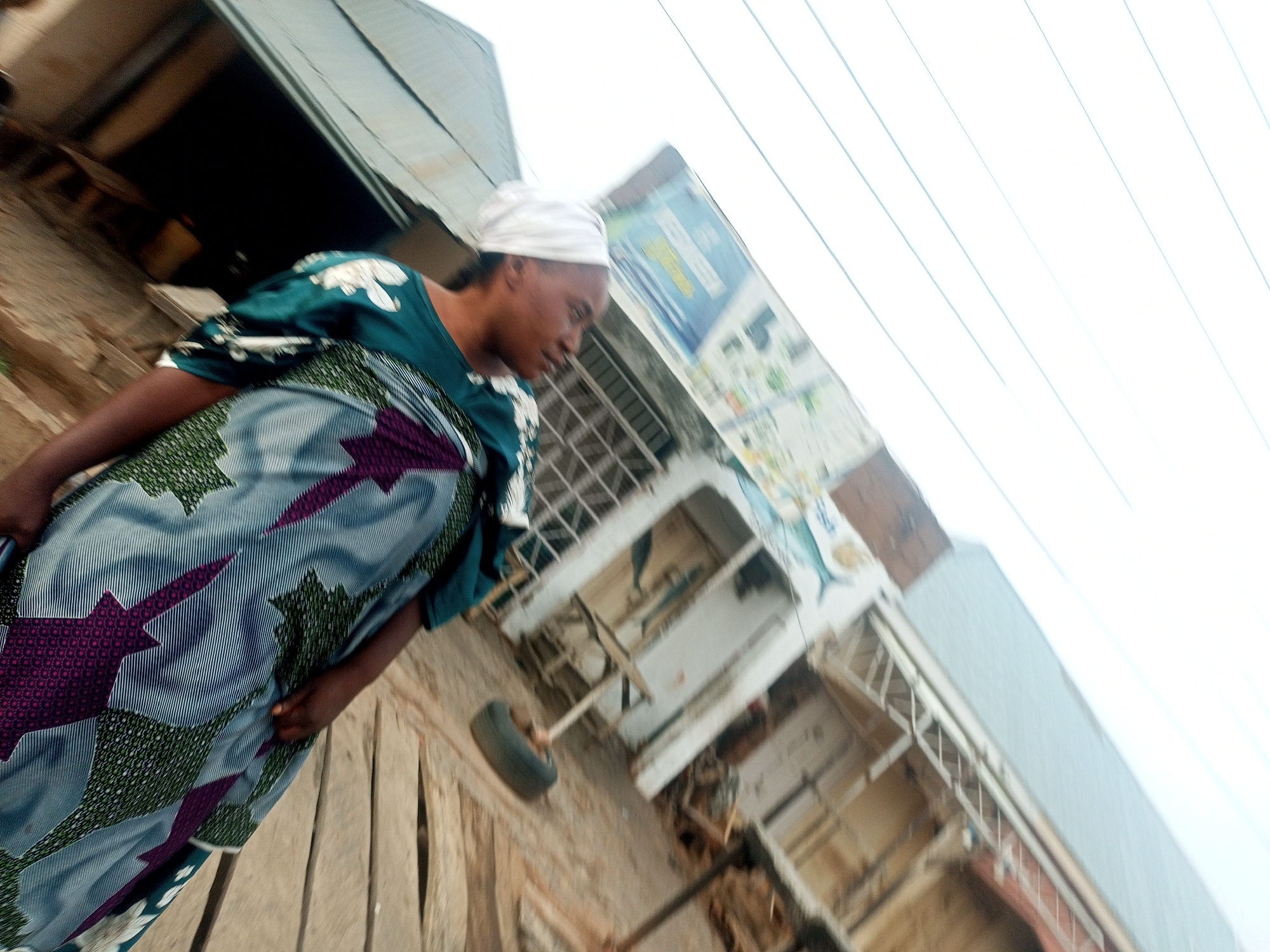 A woman wearing a white headscarf, a green and white floral blouse, and a blue skirt with purple and green star patterns is walking on a wooden boardwalk in Tempure, Nigeria. The photograph is taken from a low angle, looking up at the woman. The background includes a blurred view of buildings and electrical wires.