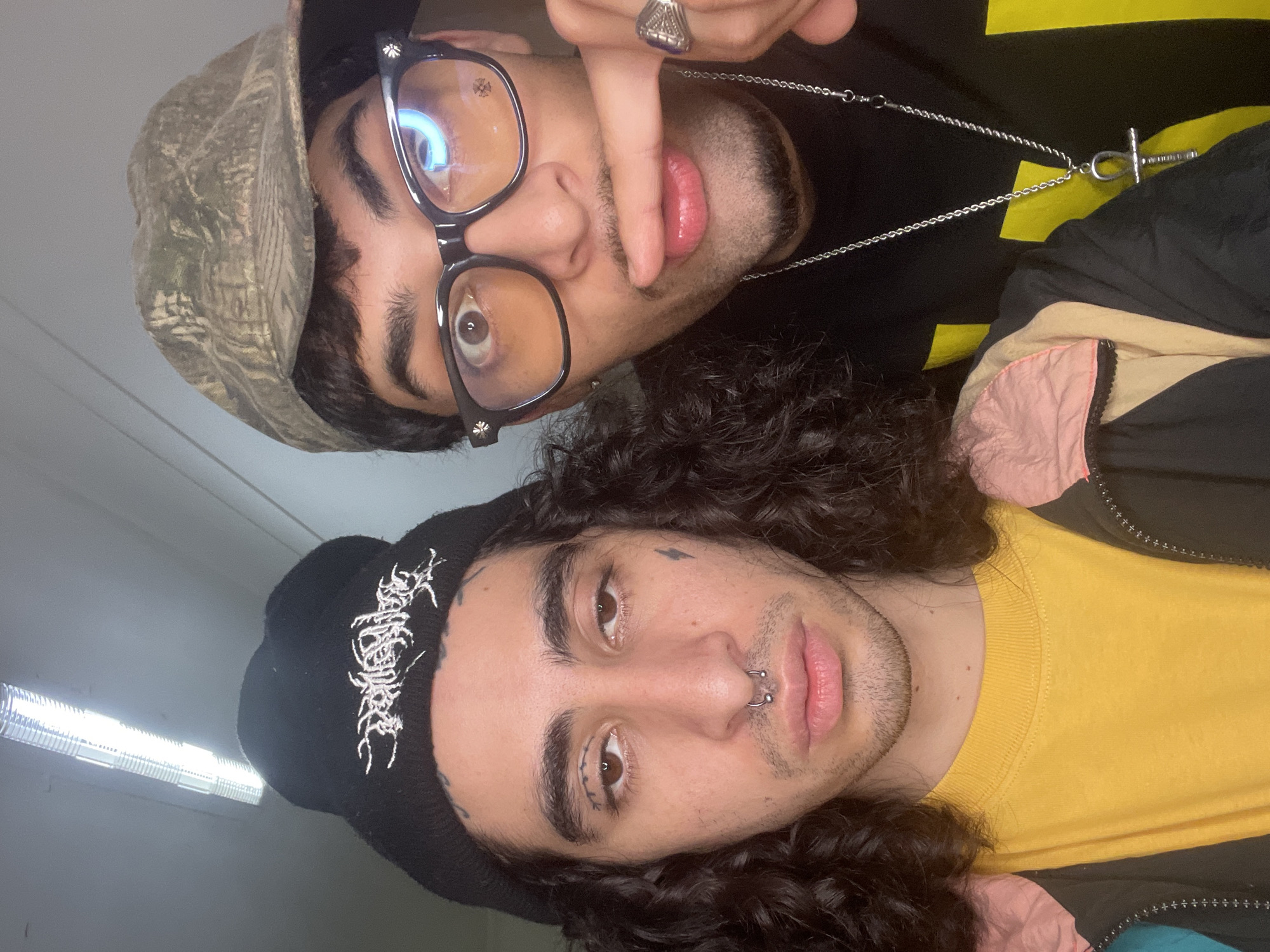 Two young men are posing for a selfie. The top figure is wearing a camouflage hat and black-framed glasses with decorative crosses. He has a goatee and a silver ring on his finger. He is wearing a black shirt with yellow stripes. He is also wearing a silver chain with an Ankh pendant. The bottom figure is wearing a black beanie with white lettering, and a nose ring. He has tattoos near his right eye and on his forehead. He is wearing a yellow shirt and what appears to be a black jacket with pink accents. The background appears to be a wall with a fluorescent light fixture. The location is unknown and there is no information to suggest the photo was taken in the city of Buenos Aires, Argentina.