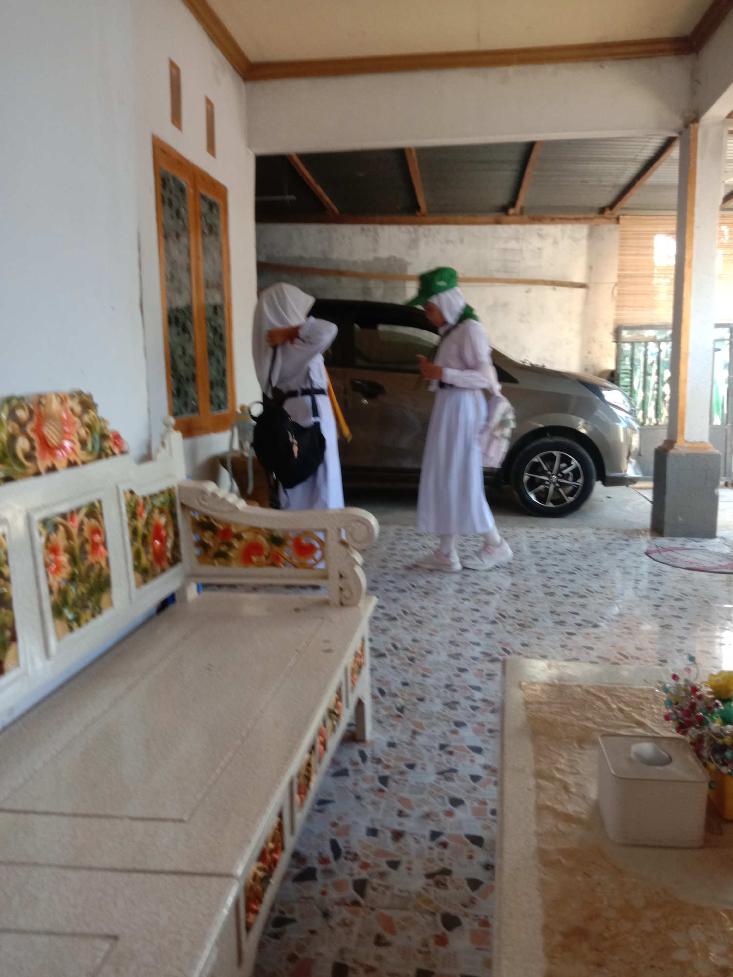 Two young girls in white hijabs and long skirts stand in what appears to be the covered outdoor area of a building. One girl, wearing a green headscarf and a green baseball cap, is in the foreground, facing away from the camera. The other girl, wearing a plain white headscarf, is further back, facing a silver car. She is holding a black bag over her shoulder and appears to be looking at her phone. A long, ornate white bench with colorful floral carvings is in the foreground, facing left. The floor is a mosaic of colorful tiles. The background shows a silver car parked under a corrugated metal roof supported by wooden beams. The scene is likely in Kwandang, Indonesia.