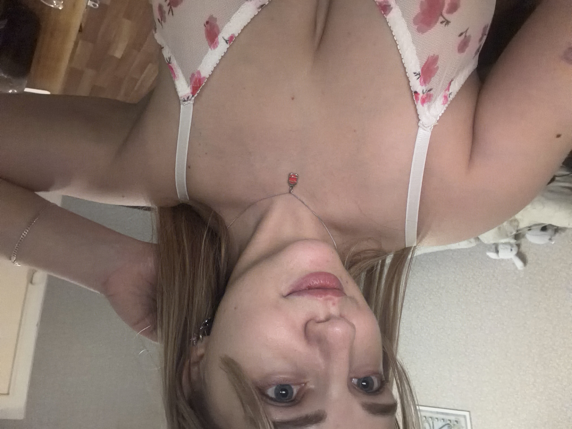 A medium shot of a light-skinned woman with shoulder-length blond hair, wearing a floral-patterned bra and a necklace with a small pendant. She is holding her left hand near her head, with a bracelet visible on her wrist. Her eyes are blue, and she is looking directly at the camera. The background is blurred, but appears to show part of a room with a wooden surface, a wall, and some objects. The location is not discernible.