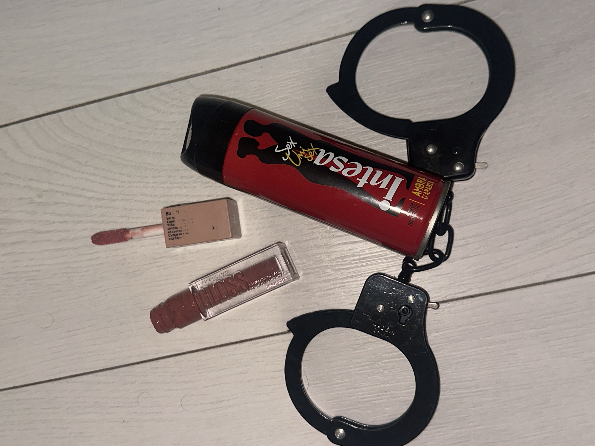 The scene depicts a collection of personal care items and accessories arranged on a light-colored wooden surface, consistent with flooring or a tabletop, within the city of Sofia, Bulgaria. The primary objects include a pair of black, metal handcuffs with an attached chain, positioned in the lower quadrants of the image. Intertwined with the handcuffs is a red aerosol can labeled "Intesa Sex Unisex PARFUM DEODORANT AMBRA D'ARABIA," featuring a black silhouette graphic of two embracing figures. To the upper left of the deodorant can, a clear rectangular bottle of "MAYBELLINE GLOSS HYALURONIC ACID" is present, uncapped with its applicator wand detached and displaying a brownish-pink liquid product. To the upper right, a smaller, square-shaped, light-beige container of what appears to be another liquid lip product is also depicted uncapped, with its applicator wand partially extended, showing a similar colored product on its tip. No individuals or active interactions are visible in the image.