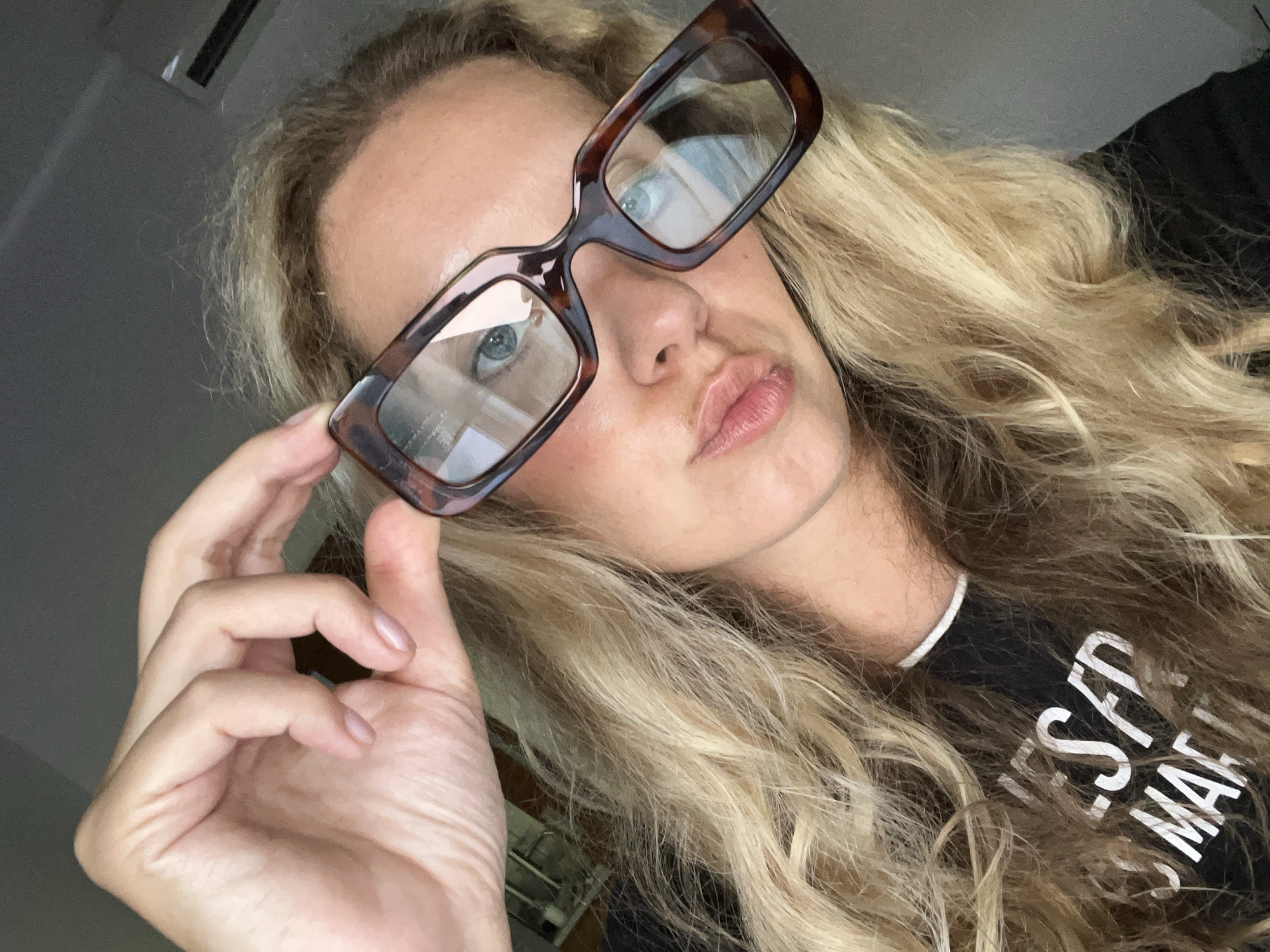 A close-up selfie shows a woman with long, wavy blonde hair and blue eyes wearing large, tortoiseshell-patterned eyeglasses. She is holding the right temple of her glasses with her left hand, her fingers gracefully positioned. Her lips are slightly pursed, creating a playful expression. She is wearing a black t-shirt with white text on it that is partially visible. The text appears to read "ESF MA". The lighting suggests it might be daytime, possibly indoors due to the lighting and background, though no specific location cues are evident beyond the clothing. The overall mood of the image is casual and self-aware. The background is dark and out of focus, with hints of what might be a ceiling fixture or a wall.