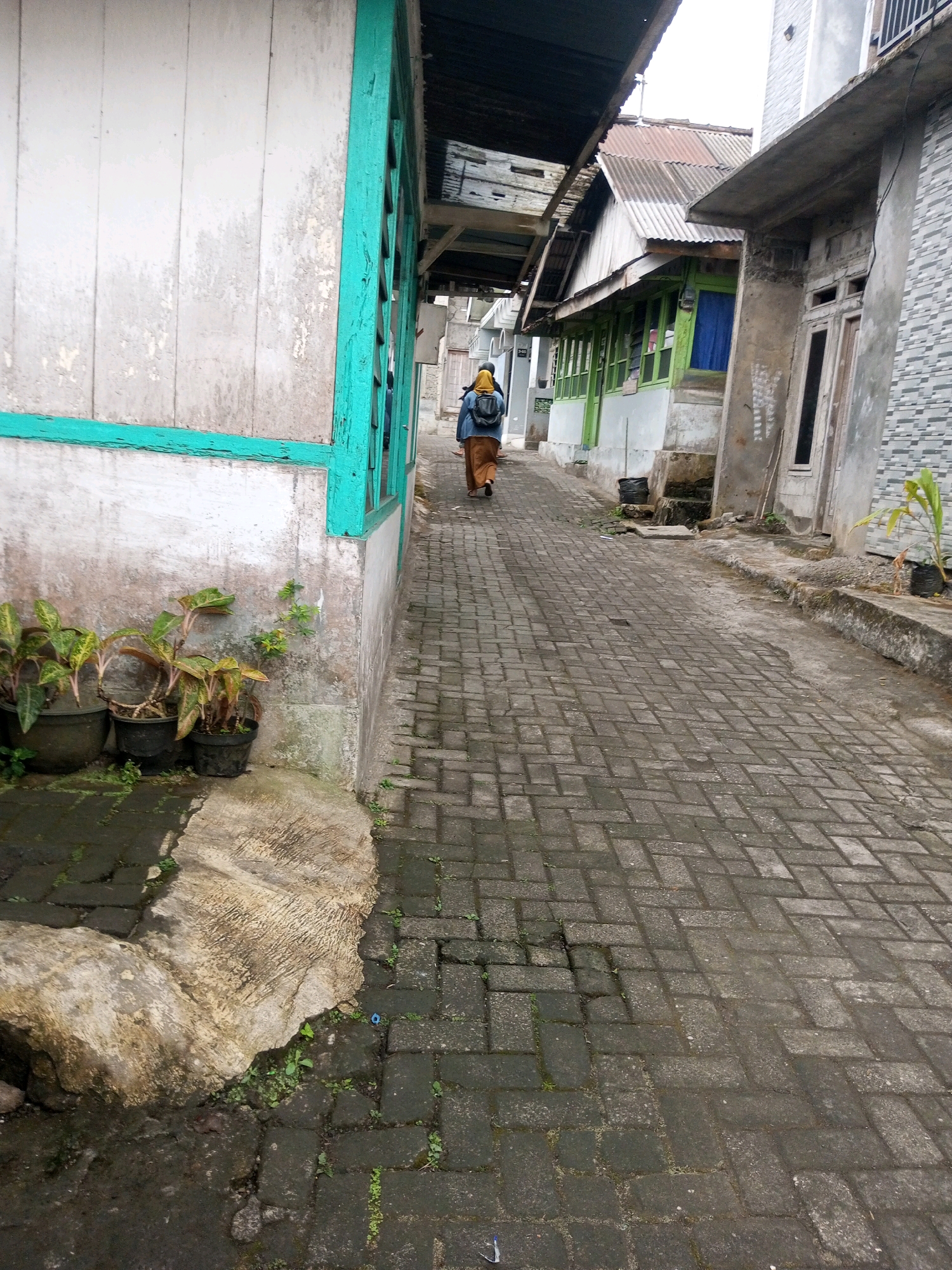 The image captures a narrow, paved alleyway in Andongsili, Indonesia. The alley is lined with buildings on both sides, featuring a mix of architectural styles. On the left, a weathered white wooden structure with turquoise trim is prominent, with several potted plants placed at its base. The alley itself is paved with dark gray interlocking bricks laid in a herringbone pattern, showing some signs of wear and moss growth. Further down the alley, on the right, a person wearing a yellow headscarf and a brown outfit with a backpack is walking away from the camera. Adjacent to this person are buildings with corrugated metal roofs and green window frames. The overall scene suggests a residential or semi-commercial area within the city, characterized by its compact development and utilitarian construction.