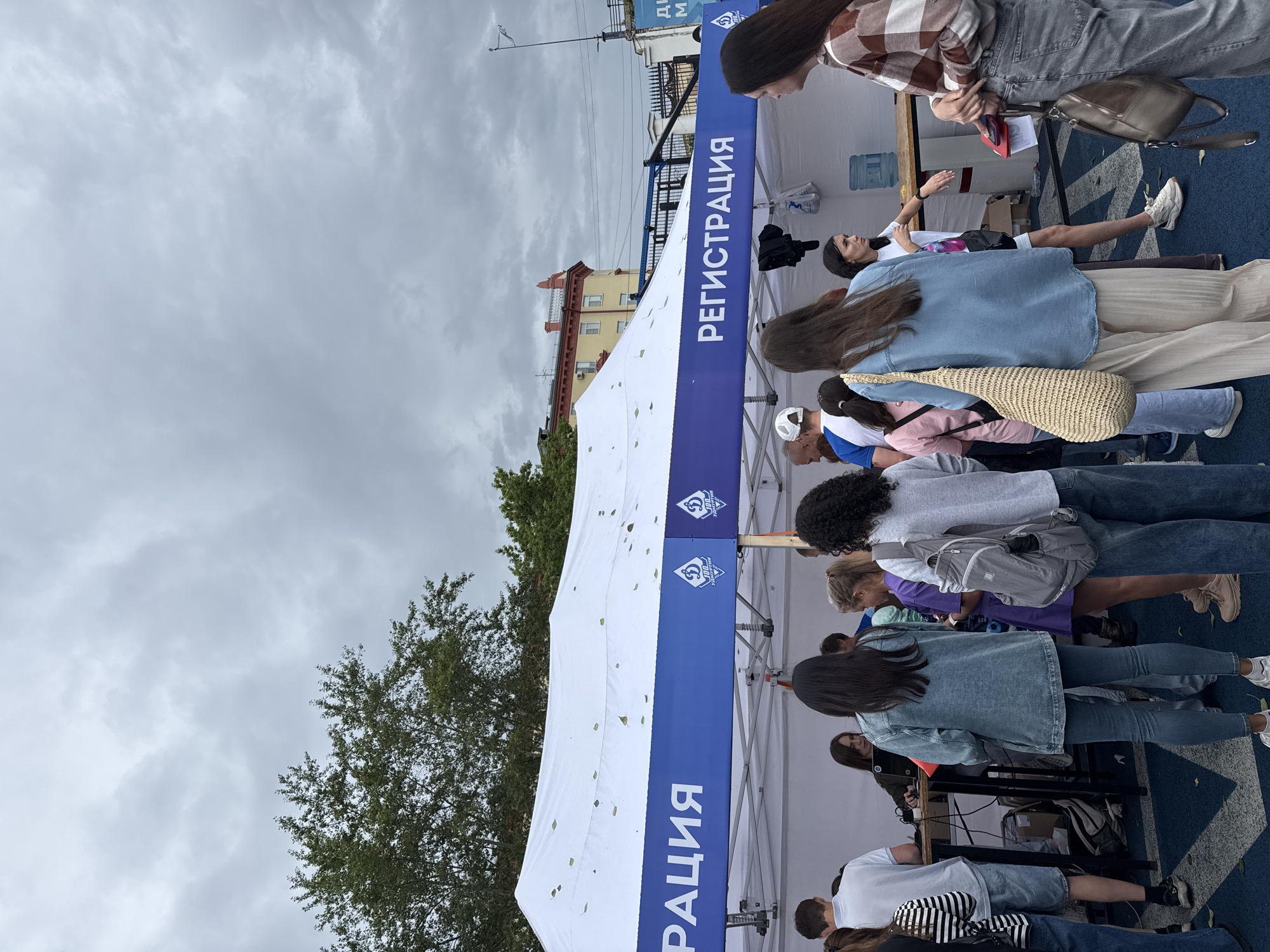 The image depicts an outdoor registration area under a white tent with a blue banner. The banner displays Cyrillic text that reads "РЕГИСТРАЦИЯ" (Registration) on both the left and right sides, as well as logos. Several people are gathered around a table under the tent, some appearing to be registering or interacting with those managing the registration.
In the background, there are trees and buildings, suggesting an urban environment. The sky is overcast with gray clouds. The ground is a dark blue surface with a geometric pattern.
The overall scene appears to be a public event or gathering where people are signing up for something. The presence of logos and the organized setup under the tent indicate an official event. The style of dress of the people suggests it is a casual daytime event.