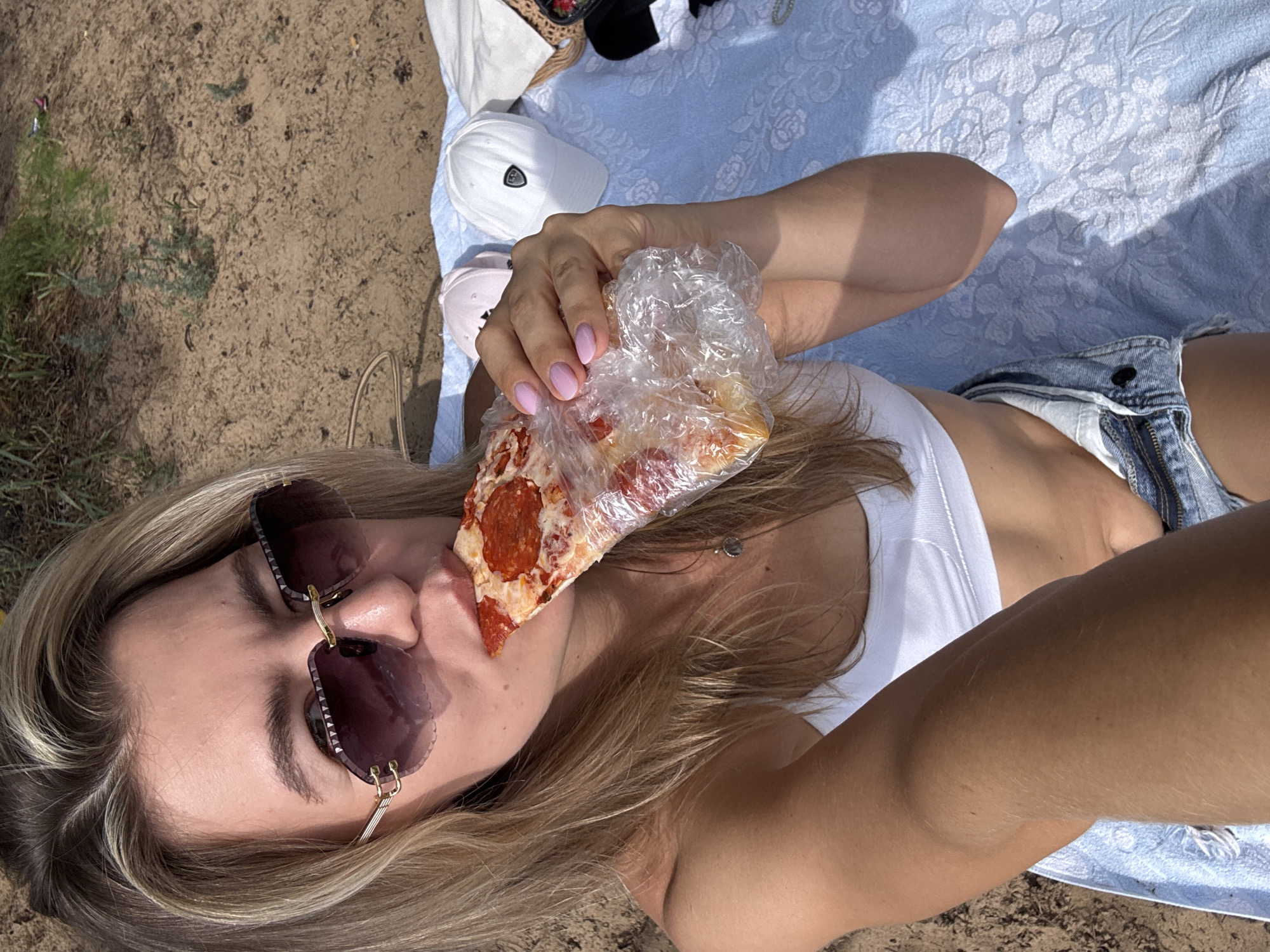 A person lies on a blue patterned blanket outdoors, with sand and some greenery visible in the background. They are wearing a white top and denim shorts, and large sunglasses. The person is holding a slice of pepperoni pizza wrapped in clear plastic and bringing it towards their mouth, with their eyes closed. Their fingernails are painted a light pink color. Several hats are visible on the blanket behind the person, including a white baseball cap and a pink cap. The lighting suggests it is daytime, with shadows indicating a sunny environment. The overall impression is one of casual relaxation and enjoying a meal outdoors. There is no visible text in the image.