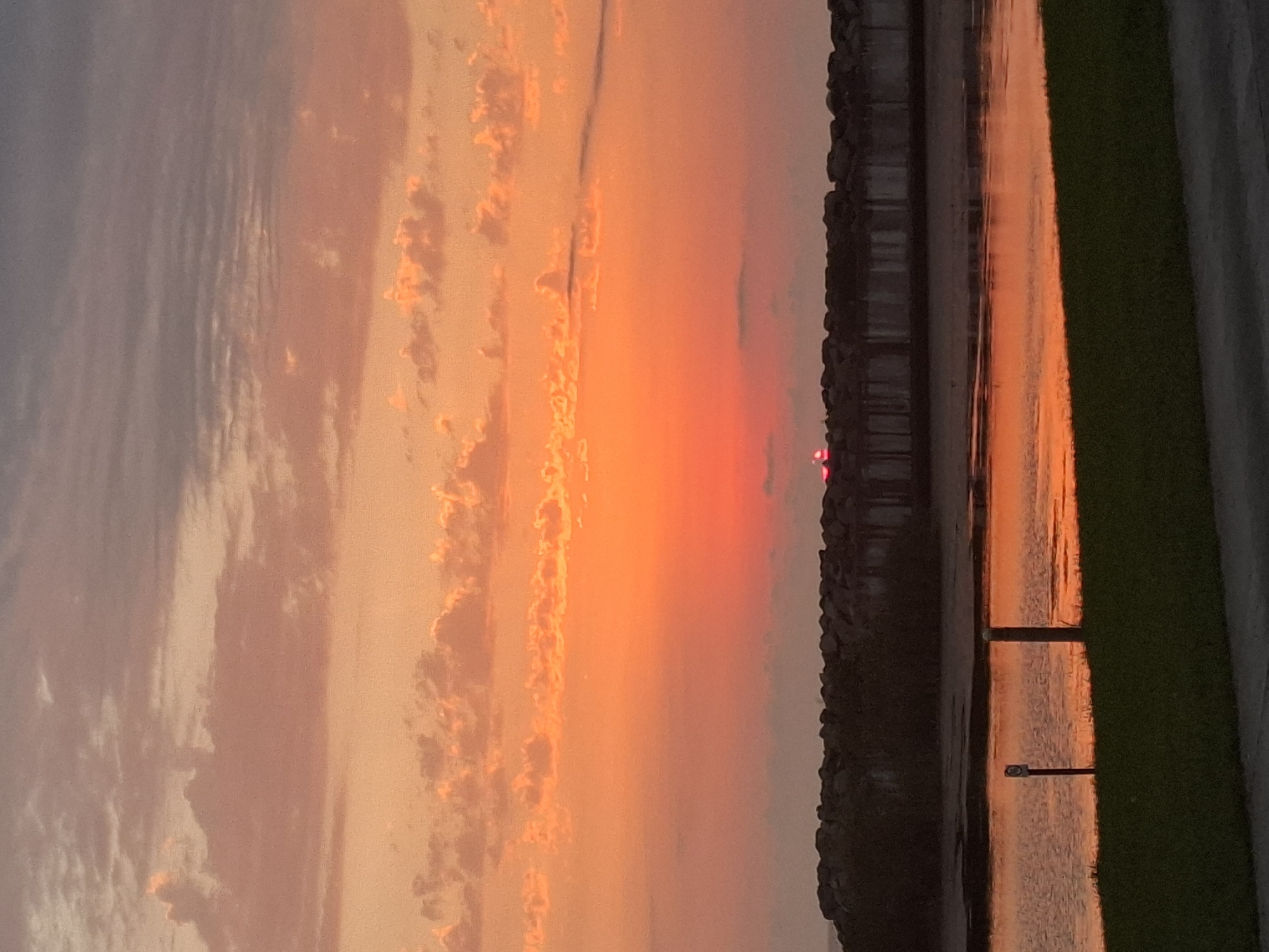 This is a photograph of a sunset over the Black Sea in Anaklia, Georgia. The sky is filled with vibrant orange and pink clouds, and the sun is setting on the horizon, casting a warm glow on the water. In the foreground, there is a grassy area and a paved path. A breakwater made of large rocks extends into the water, and behind it, structures that appear to be part of a boardwalk or pier can be seen. The water reflects the colors of the sky, creating a beautiful and tranquil scene. There are no people visible in the image.