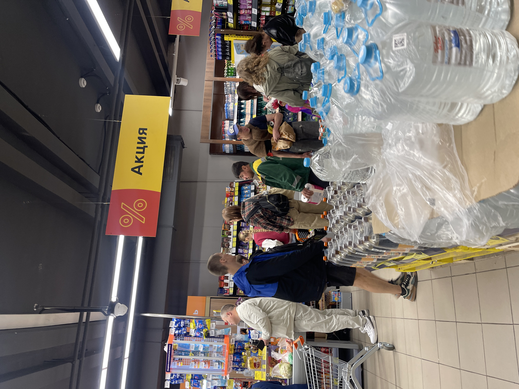 The image depicts a scene inside a supermarket in Moscow, Russia. Several people are present, browsing or shopping. In the foreground, a large display of bottled water is visible. The shelves in the background are stocked with various products. Signs with percentages and the Russian word "Акция" (which translates to "Sale" or "Promotion") are hanging from the ceiling, indicating a sale event. The overall environment suggests a busy retail setting.