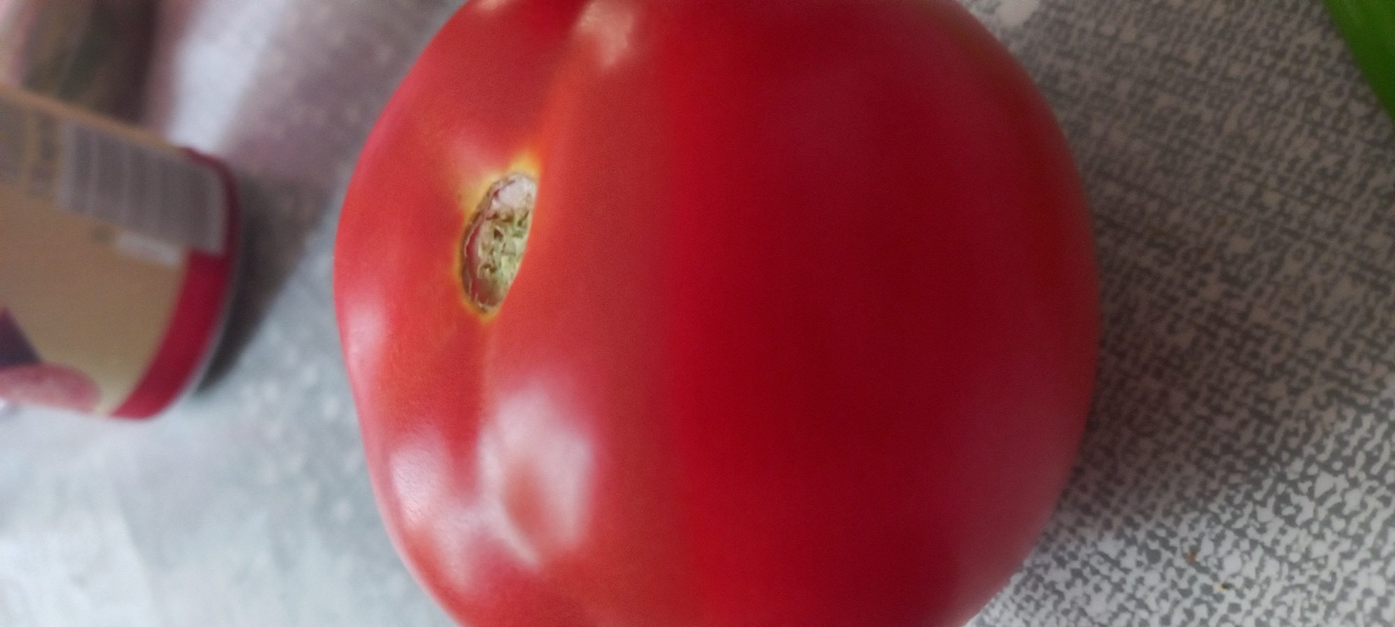 A single, ripe, red tomato is positioned prominently in the foreground, occupying the lower and central portions of the frame. Its surface is a uniform deep red, exhibiting subtle reflections, particularly a lighter, somewhat oval-shaped highlight on the left side of its upper half. Near the top of the tomato, a distinct indentation reveals a small cavity where the stem was previously attached, showing remnants of light green to yellowish-green fibrous material. The tomato rests on a flat, light grey surface patterned with irregular, darker grey, interlocking shapes. In the blurred background, to the upper right, a cylindrical object is partially visible. It features a light beige or tan body with a distinct red band near its base. Indistinct vertical lines, possibly text or a barcode, are faintly visible on its beige section.