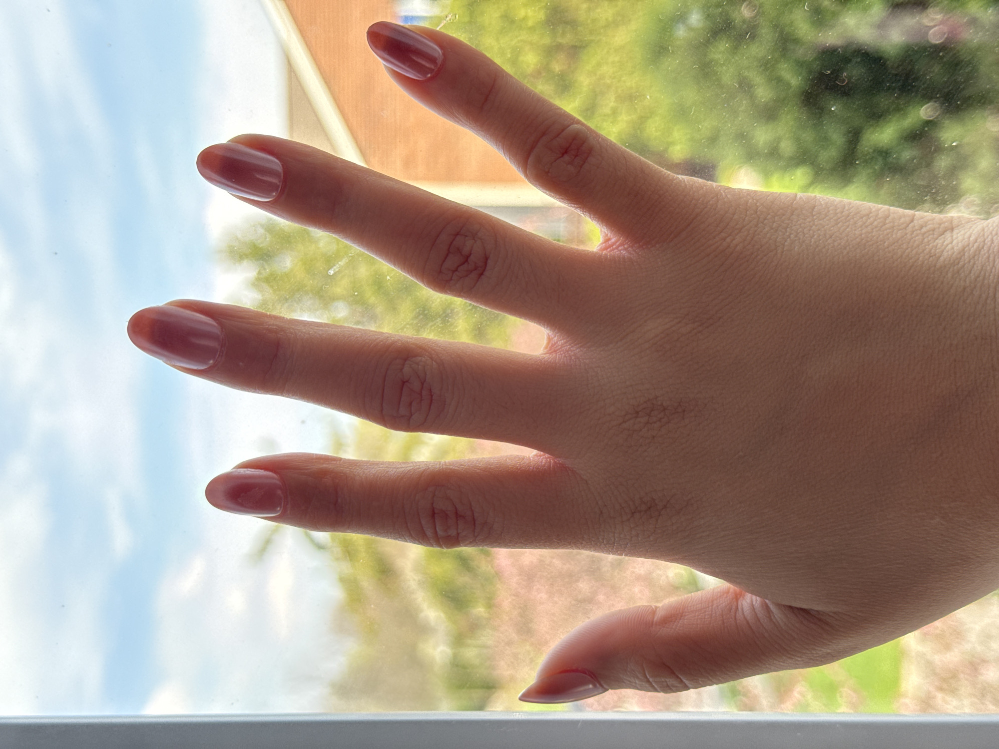 A person's hand is pressed against a window, with the fingers slightly spread. The fingernails are manicured with a glossy, semi-transparent pinkish-brown polish. Outside the window, there is a blurred view of foliage and buildings. The scene appears to be located indoors, with an exterior view, in a residential area. Without further data, it is not possible to pinpoint the exact location in the city of Columbus, Ohio, United States of America.