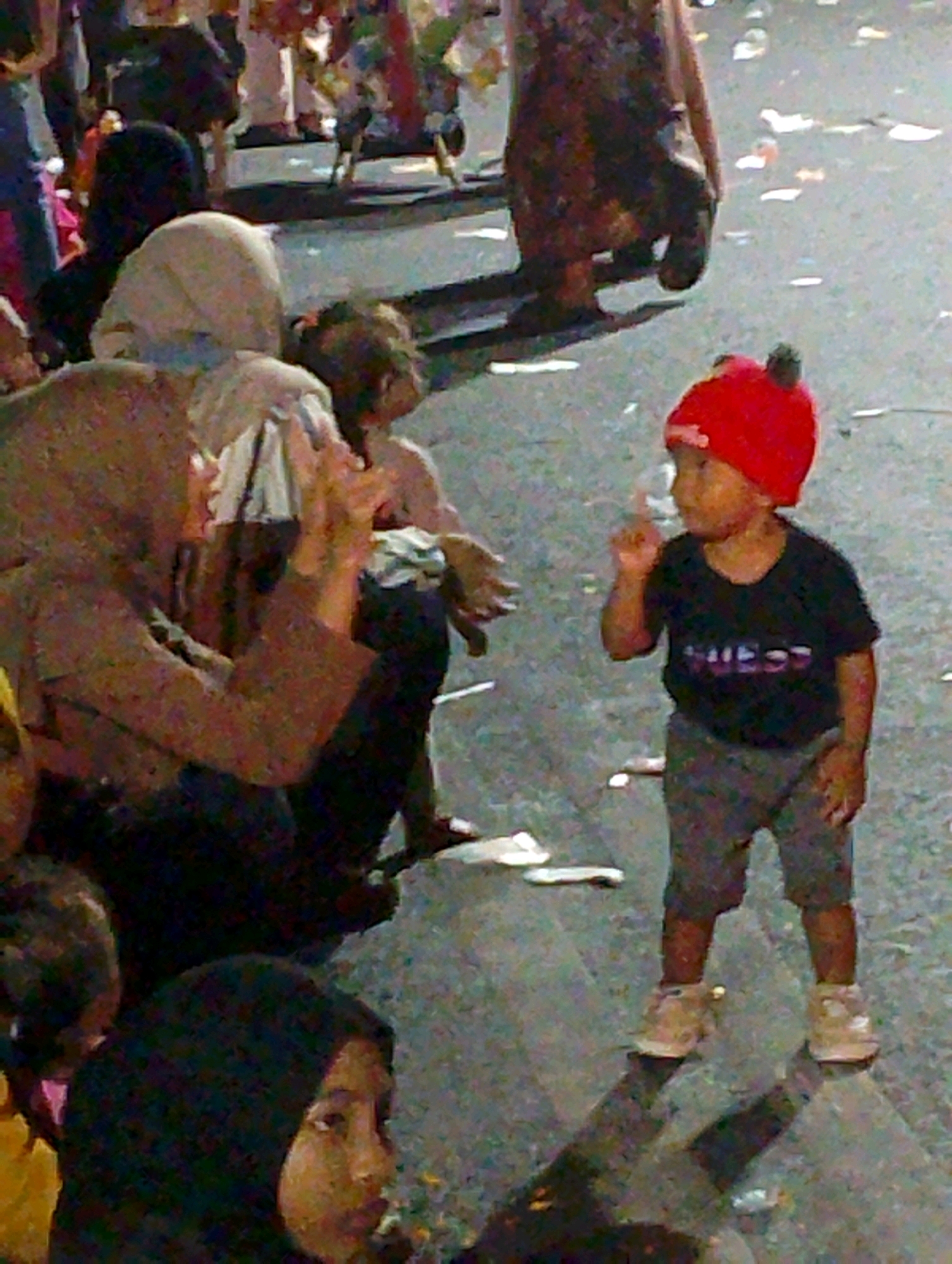 The image depicts a street scene with several people, including children, on what appears to be a paved surface. In the foreground, a young child wearing a red beanie and a black t-shirt with white text is looking to the left and has their right index finger raised. To the left of this child, a person wearing a tan headscarf is visible from the chest up, holding something white. Further back, other individuals are present, some partially obscured. The background suggests a gathering or event with people and objects that are out of focus. The ground is a dark, mottled surface with scattered white debris. This scene could be located in Probolinggo, Indonesia, given the context provided.