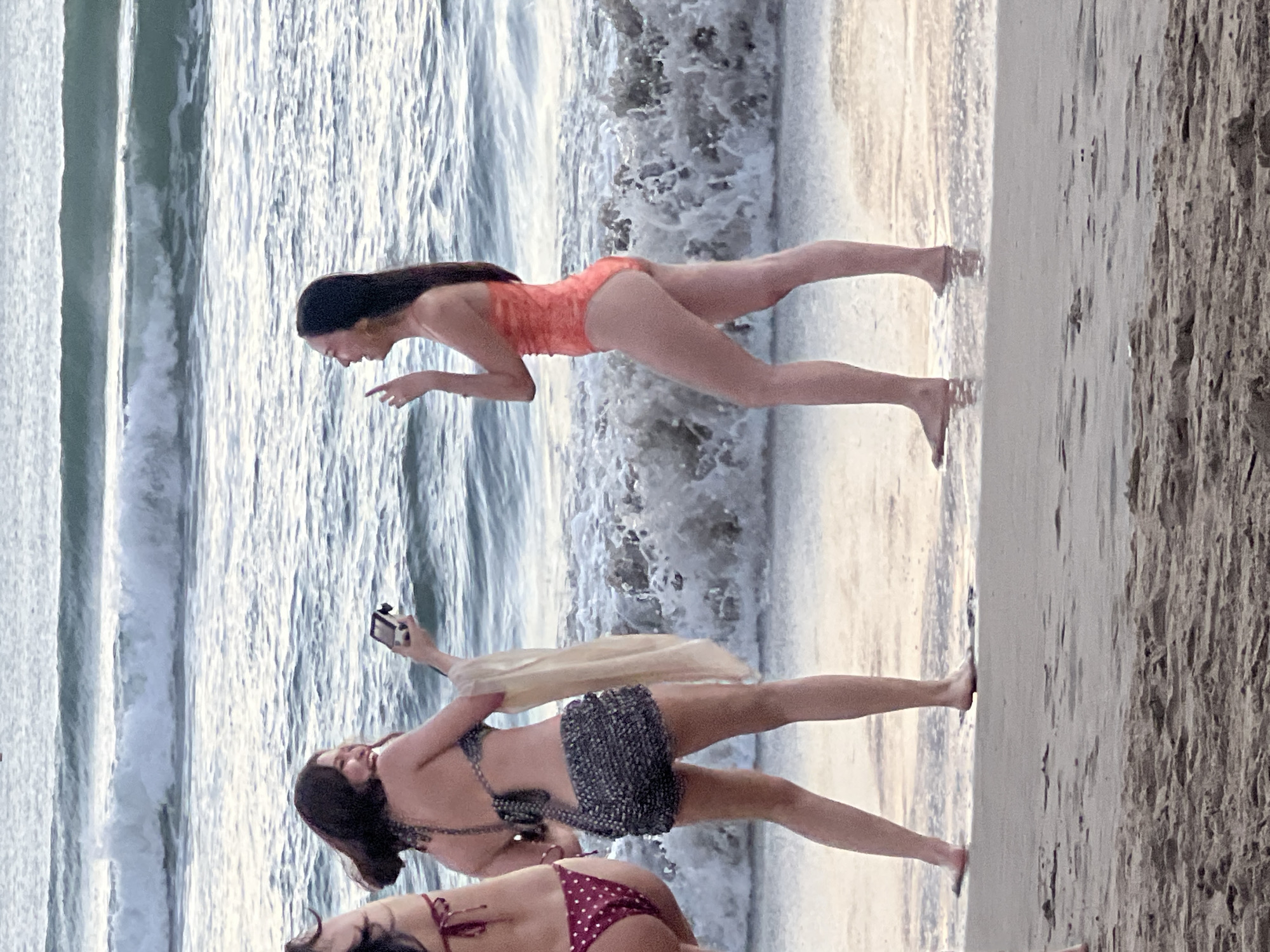 Two young women are standing on a beach in Kuta, Indonesia. The woman on the left is wearing a dark, sequined bikini top and shorts, and is holding a phone up to take a photo of the woman on the right. The woman on the right is wearing an orange one-piece swimsuit and is smiling. Both women are facing the ocean. The waves are crashing on the shore, and the sand is wet. The sky is overcast.