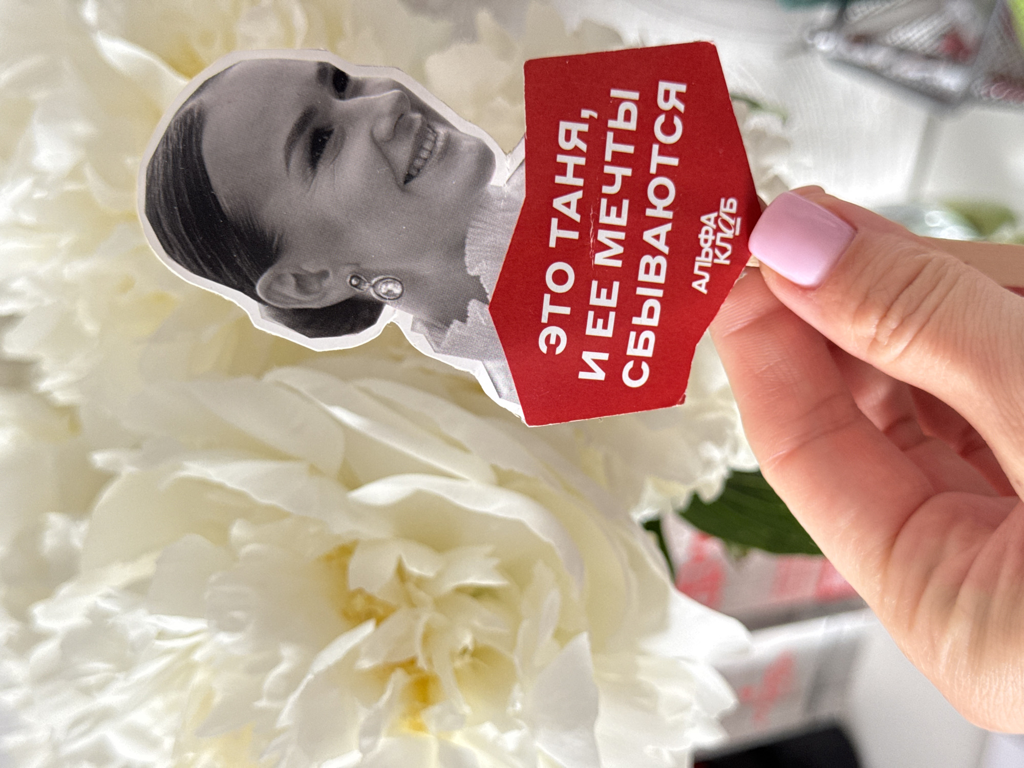 A photograph shows a hand holding a cutout image of a woman in black and white with a red card with white text on it. The text is in Russian and includes the words "Alpha Club." The background is a cluster of white flowers. The setting cannot be identified as Sochi, Russia based on the provided image.