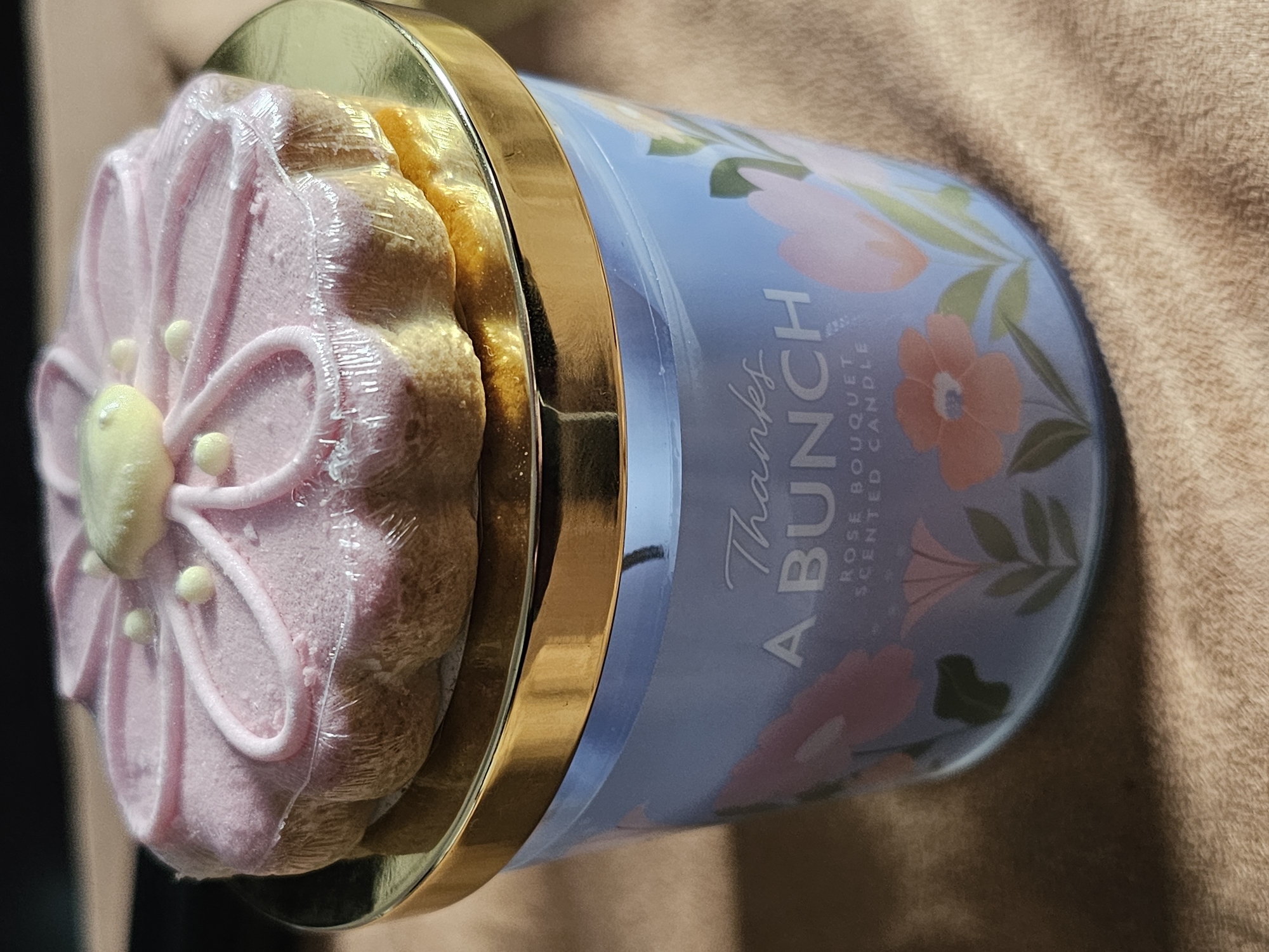 A close-up shot reveals a "Thanks A Bunch Rose Bouquet Scented Candle" with a cake-like lid wrapped in plastic. The lid features a pink floral design with a yellow center and white accents. The candle is positioned on a brown textured surface. The location is unknown.