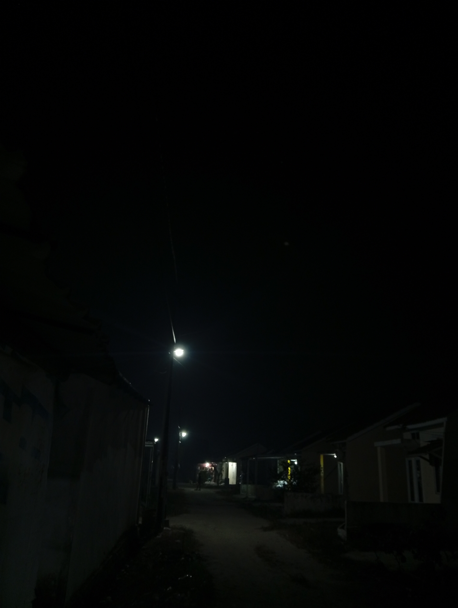 The image shows a dark, unlit street in what appears to be a residential area at night. A single streetlight illuminates a portion of the road and sidewalk. Several houses are visible on the right side of the street, with some having faint lights on inside. A person or persons are faintly visible in the distance, walking on the street. The left side of the street is dominated by a dark, imposing structure, possibly a building or wall, that obscures further detail. The sky is completely black, indicating a lack of moonlight or significant ambient light. The overall impression is one of quietness and darkness, typical of a rural or suburban street at night.