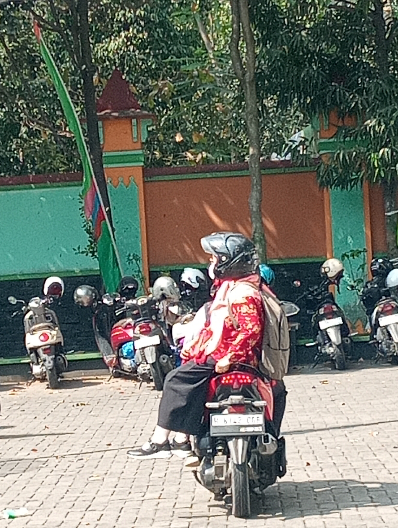 The image shows a street scene in Demak, Indonesia, with several motorcycles parked or in motion. In the foreground, a person wearing a red patterned top and a black helmet is seated on a red motorcycle, with a backpack on their back. The motorcycle has a license plate that reads "B 6142 CFE". The ground is paved with interlocking bricks. In the background, there is a building with a teal and orange facade, and trees with green foliage. Other motorcycles, some with riders wearing helmets, are visible. A green flag is also visible, attached to a pole. The lighting suggests it is daytime, with shadows cast on the ground.