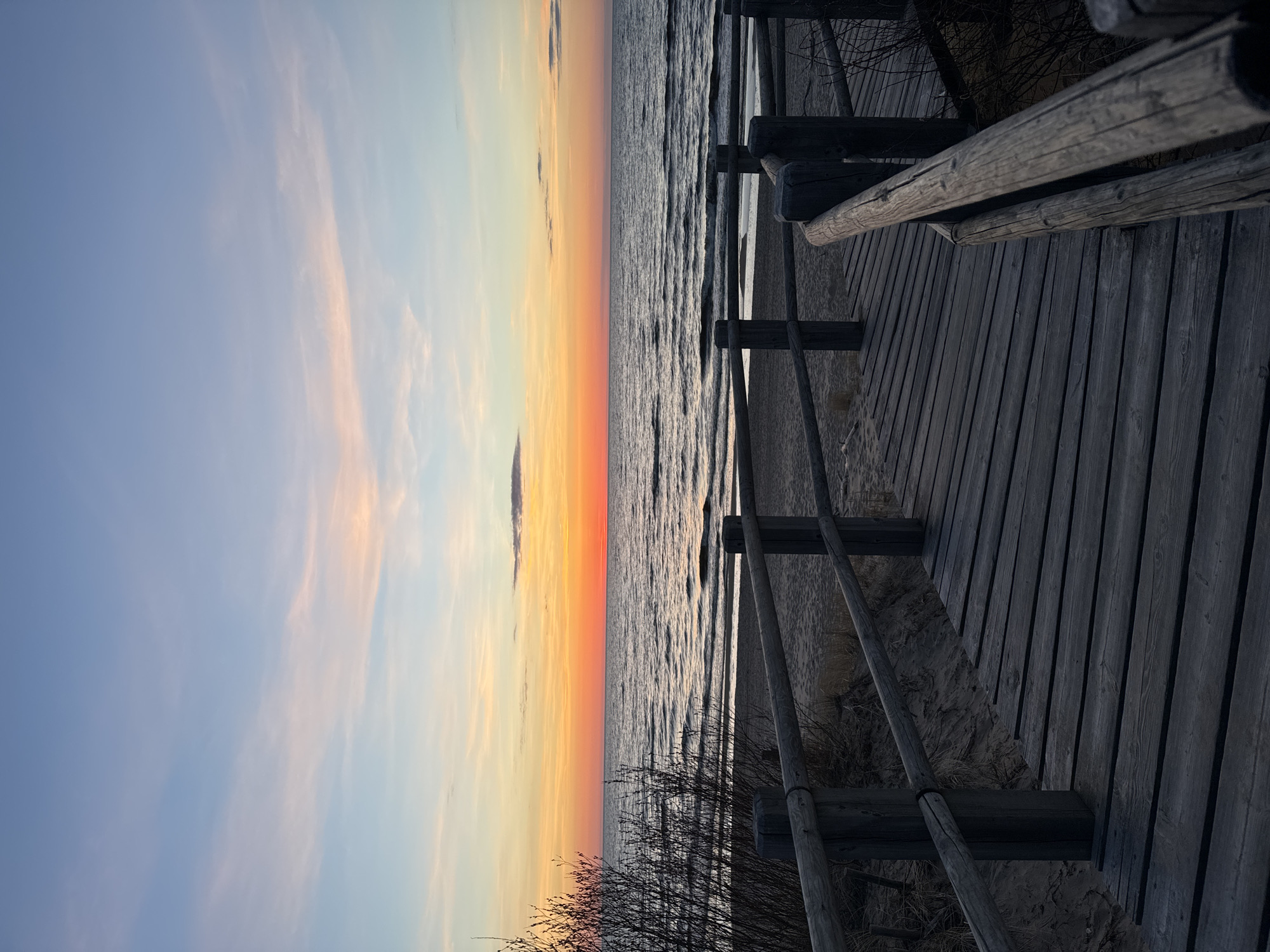 The image depicts a sunset view at a beach in Riga, Latvia. The scene is oriented vertically.
On the right side of the image is a wooden boardwalk with a railing. The boardwalk runs along the beach. In the foreground, there are some dry bushes growing near the boardwalk.
The beach extends to the center of the image, where the water of the Baltic Sea meets the sky. The sky displays a gradient of colors, from blue on the left, transitioning to yellow and orange in the center, indicating a sunset. There are some light clouds visible in the sky. The water is slightly choppy with small waves.