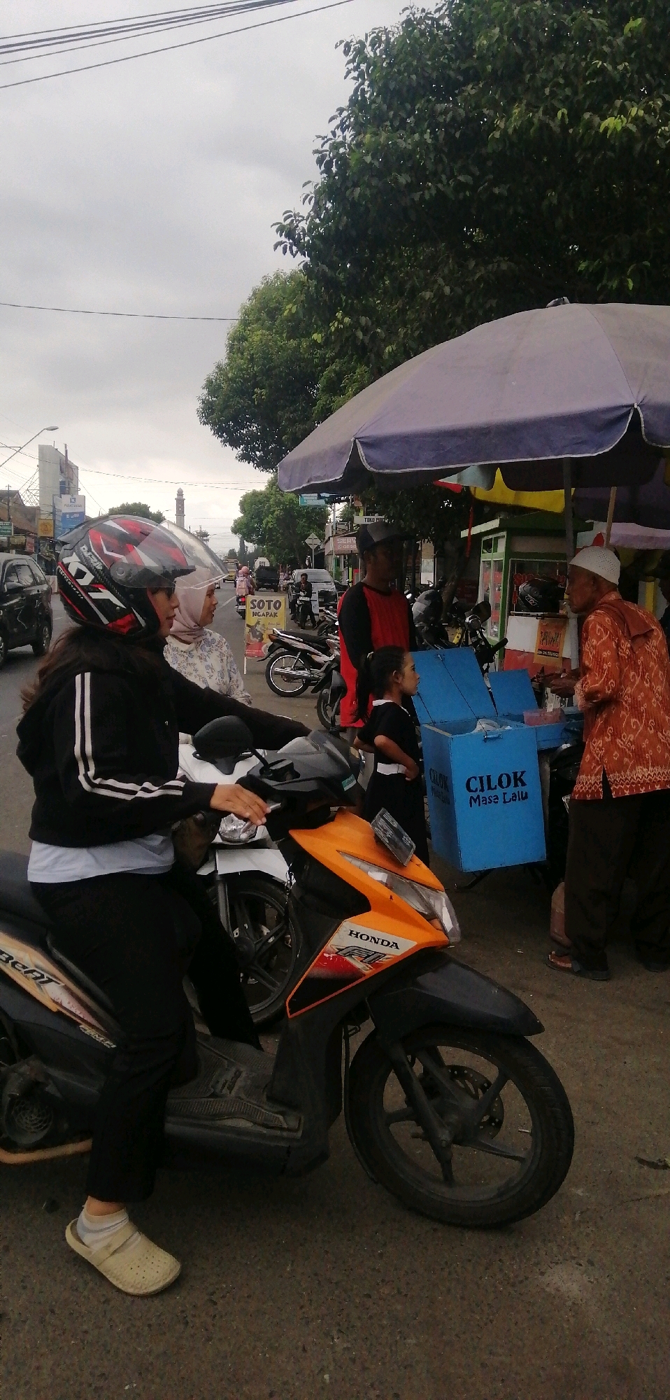 A roadside scene in Ajibarang, Indonesia, depicts an outdoor food stall under a large umbrella. A woman wearing a black jacket, white shirt, black pants, and a red and black helmet is seated on an orange and black Honda scooter. She is looking towards the right side of the frame. To her right, a woman wearing a patterned white and blue shirt and a light pink hijab stands looking towards the food stall. Further to the right, a child in a black shirt and skirt stands by a blue cooler box labeled "CILOK Masa Lalu". An elderly man in an orange patterned shirt and white cap is interacting with someone behind the cooler. Another man in a red and black shirt stands between the child and the elderly man. The environment includes a street with parked motorcycles, a building with signage that includes "SOTO NGAPAK", and trees lining the background. The sky is overcast.