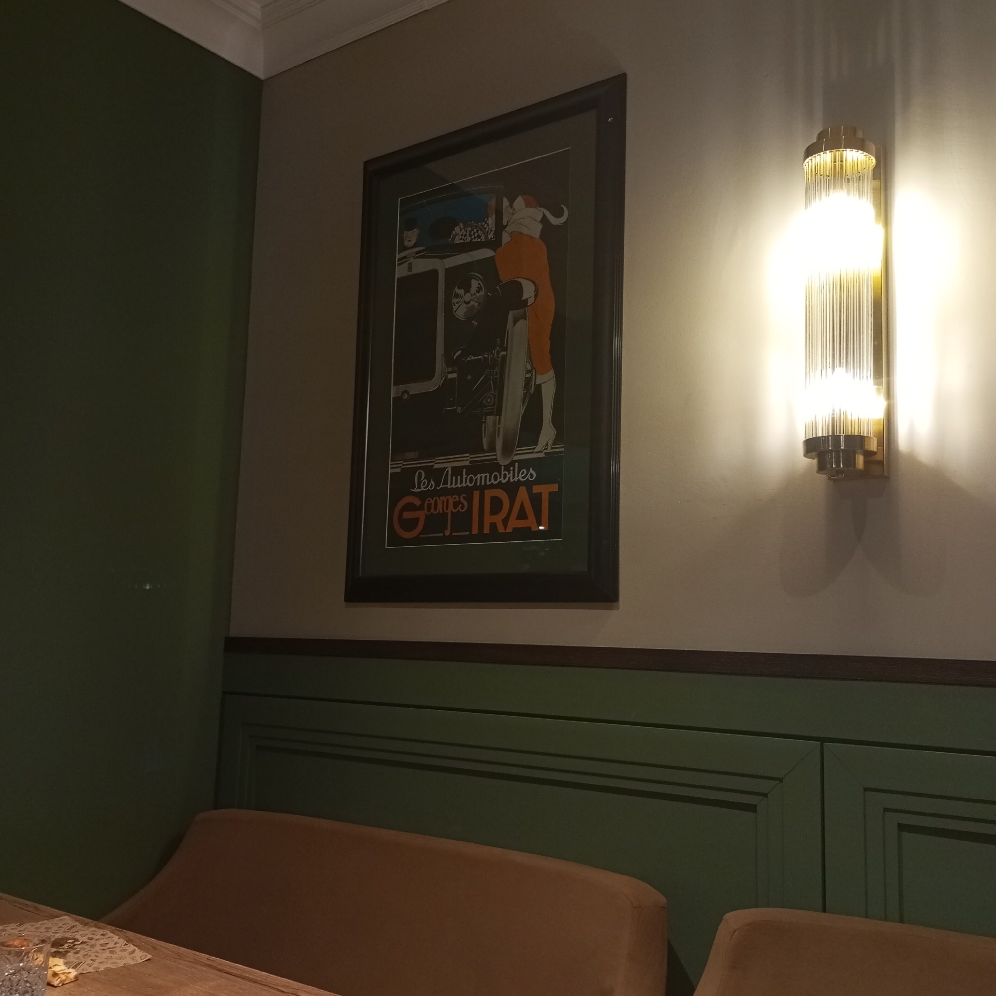 The image shows an interior scene with dark green walls and light beige walls, separated by a dark brown wooden trim. On the light beige wall hangs a framed vintage advertisement poster depicting a woman in an orange jumpsuit driving a car. To the right of the poster is a modern wall sconce with vertical glass tubes that emits a bright, warm light, casting shadows on the wall. In the foreground, a wooden table with a patterned placemat and a glass is visible, along with a portion of a light brown upholstered bench or sofa. The scene appears to be in a restaurant or bar. The location is not specified to be in Izhevsk, Russia.
