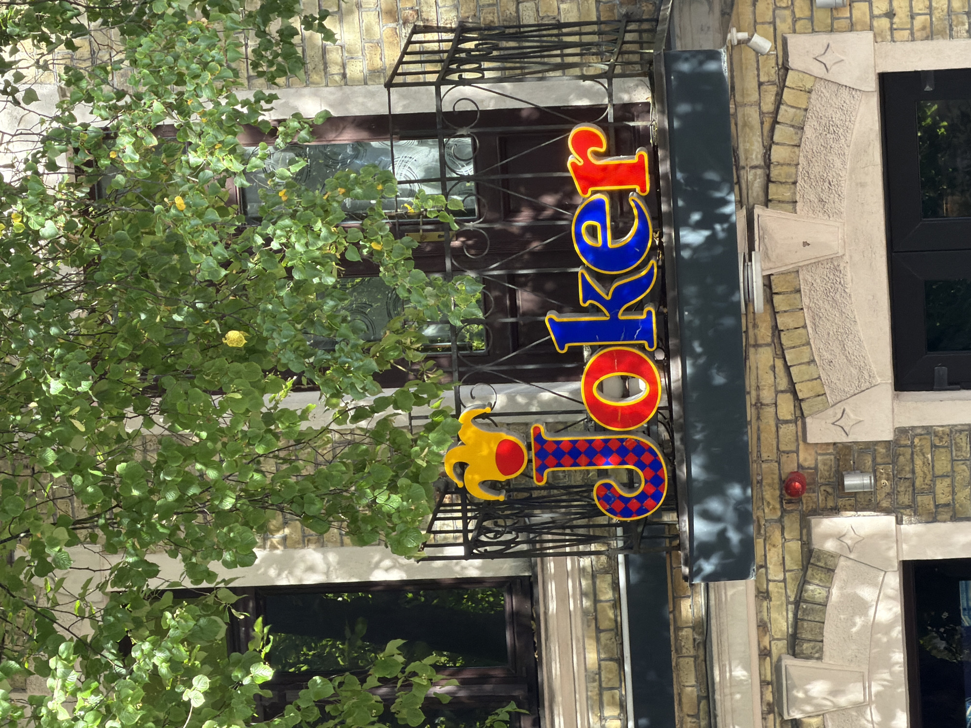 The image shows a close-up view of a sign that says "Joker" in large, colorful letters. The letters are red and blue with yellow outlining. Above the letters is a yellow jester's hat with red accents. The sign is mounted on a balcony railing in front of a brick building. A tree with green leaves partially obscures the sign. The lower part of the image shows architectural details of the building, including arched doorways and decorative stonework. Based on the architecture and signage, this appears to be a commercial establishment, likely a pub or entertainment venue.
The "Joker" sign is located in Ventspils, Latvia.