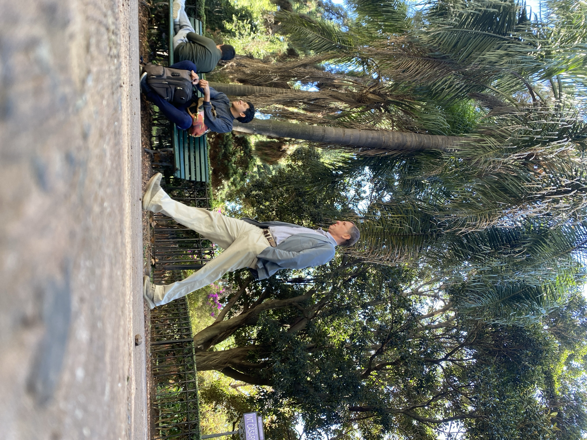 The image shows a sunny outdoor scene in Buenos Aires, Argentina, likely within the Parque Lezama. In the foreground, a man stands posing on a walkway, wearing a light blue blazer, button-down shirt, light-colored pants, and sneakers. He is turned to his right, looking up and slightly away from the camera. Behind him, a wrought iron fence separates the walkway from a grassy area with trees and flowers. In the background, two people are sitting on a green park bench. The first is sitting in the middle, facing slightly to the left with a backpack next to him. The second man is sitting on the right, dressed in a green shirt with his head and shoulders visible. The sky is visible through the trees and palm trees in the image. A sign near the bottom of the image says "INT. CASSAR".