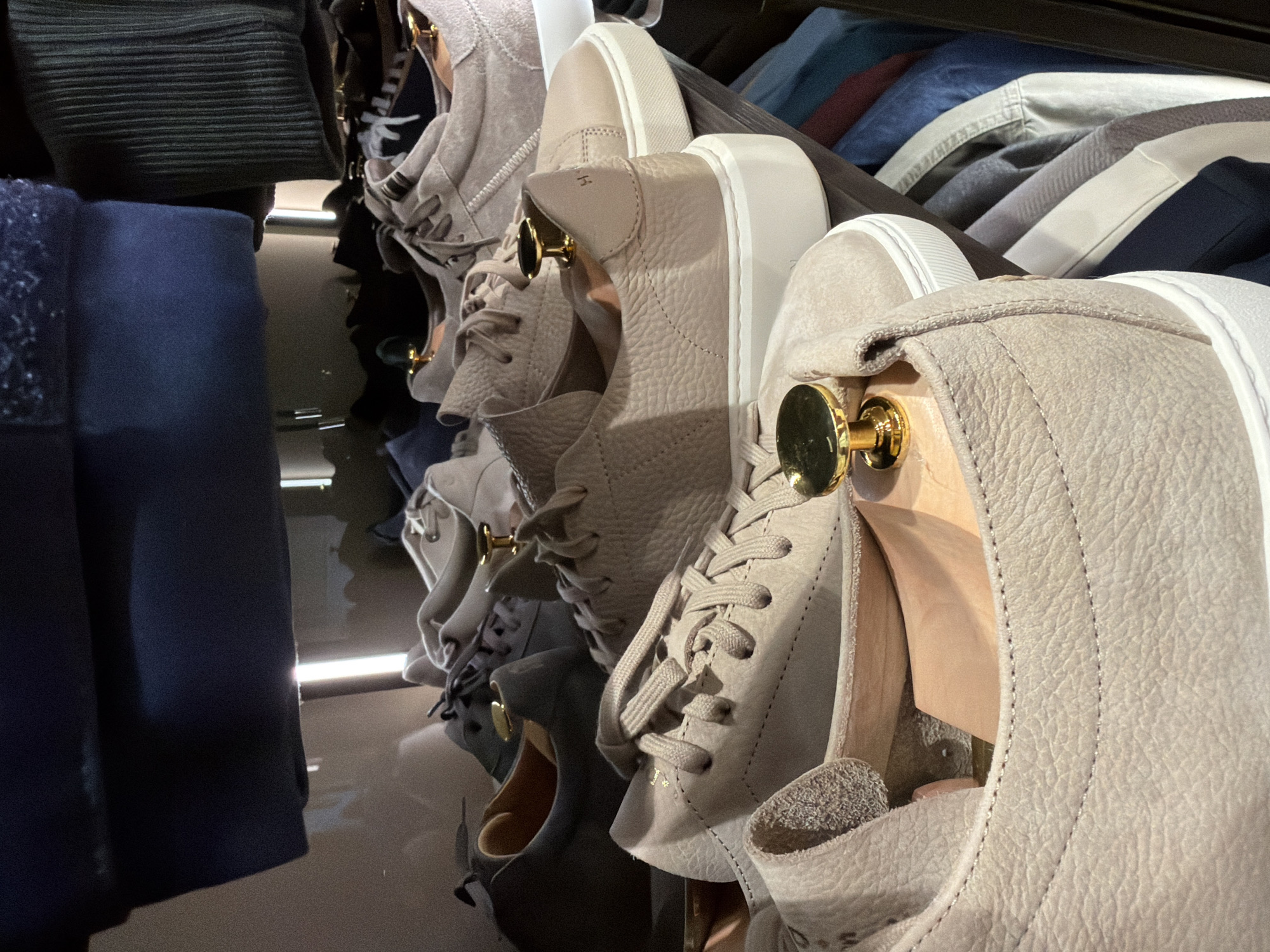 The image shows a close-up, eye-level, interior shot of a closet or retail display featuring several pairs of men's low-top sneakers. The sneakers are predominantly in neutral, light tan or beige colors, with some appearing to be made of textured leather. They are arranged on shelves, with some shoes clearly visible in the foreground and others further back. Some of the sneakers have visible laces and thick white rubber soles. Gold-colored shoe trees or formers are inserted into some of the shoes, suggesting they are new or being stored for preservation. In the background, out-of-focus clothing items, such as dark blue garments and what appears to be a ribbed knit fabric, are visible on hangers or shelves, indicating a wardrobe or retail environment. The lighting suggests an indoor setting, possibly illuminated by artificial light sources. There are no people or discernible action captured in the image. The scene does not provide any specific visual cues to identify its location in Moscow, Russia.