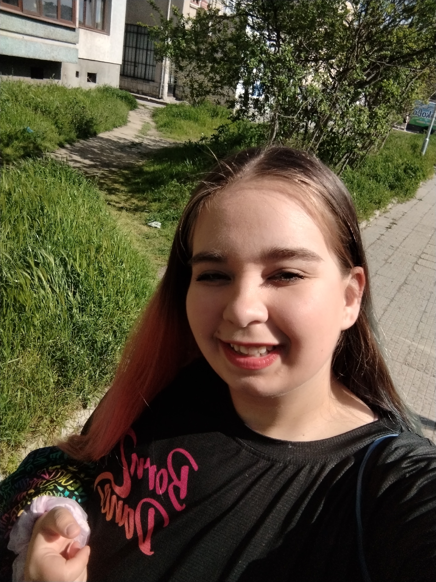 A young person is taking a selfie on a sunny day in Plovdiv, Bulgaria. The individual has fair skin, dark hair with pink highlights on one side, and is wearing a black t-shirt with a bright pink design. They are smiling at the camera and holding a scrunchie in their left hand.
The background shows a paved sidewalk, green grass, a pathway leading towards a building, and some trees. A small part of a sign is also visible. The light suggests a sunny day. The buildings appear to be older, typical of urban Plovdiv.