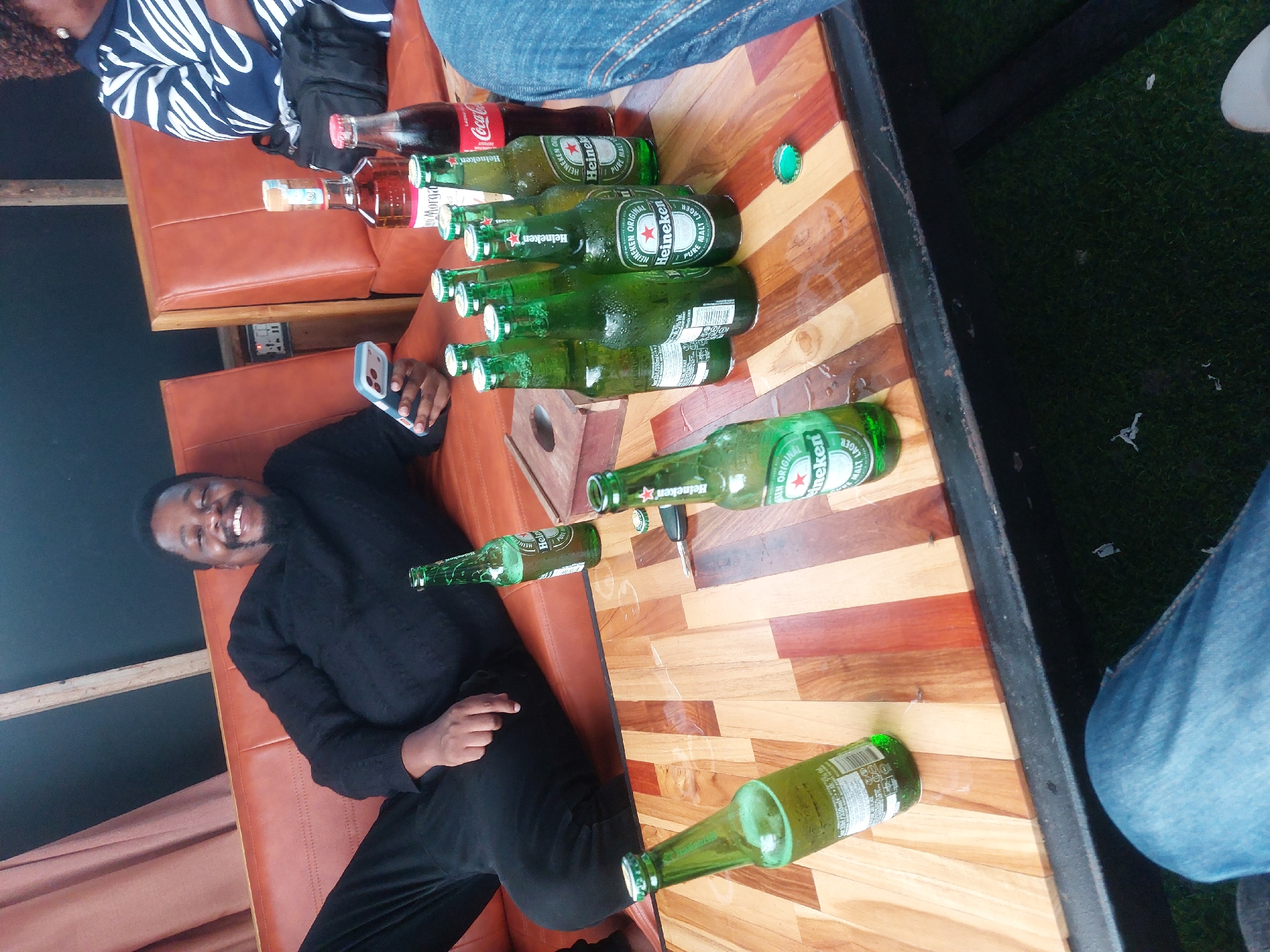 A man in a black sweater and pants lounges on an orange leather couch, holding a phone. Next to him on a wooden table are multiple empty green Heineken beer bottles, a Coca-Cola bottle, and a bottle of rum. A woman in a blue and white striped shirt sits nearby. The location appears to be an outdoor seating area, possibly a bar or restaurant, with artificial grass visible. Although not explicit in the image, given the context of the request and focus on beer bottles, it is plausible to assume this scene is set in or near Dar es-Salaam, Tanzania.