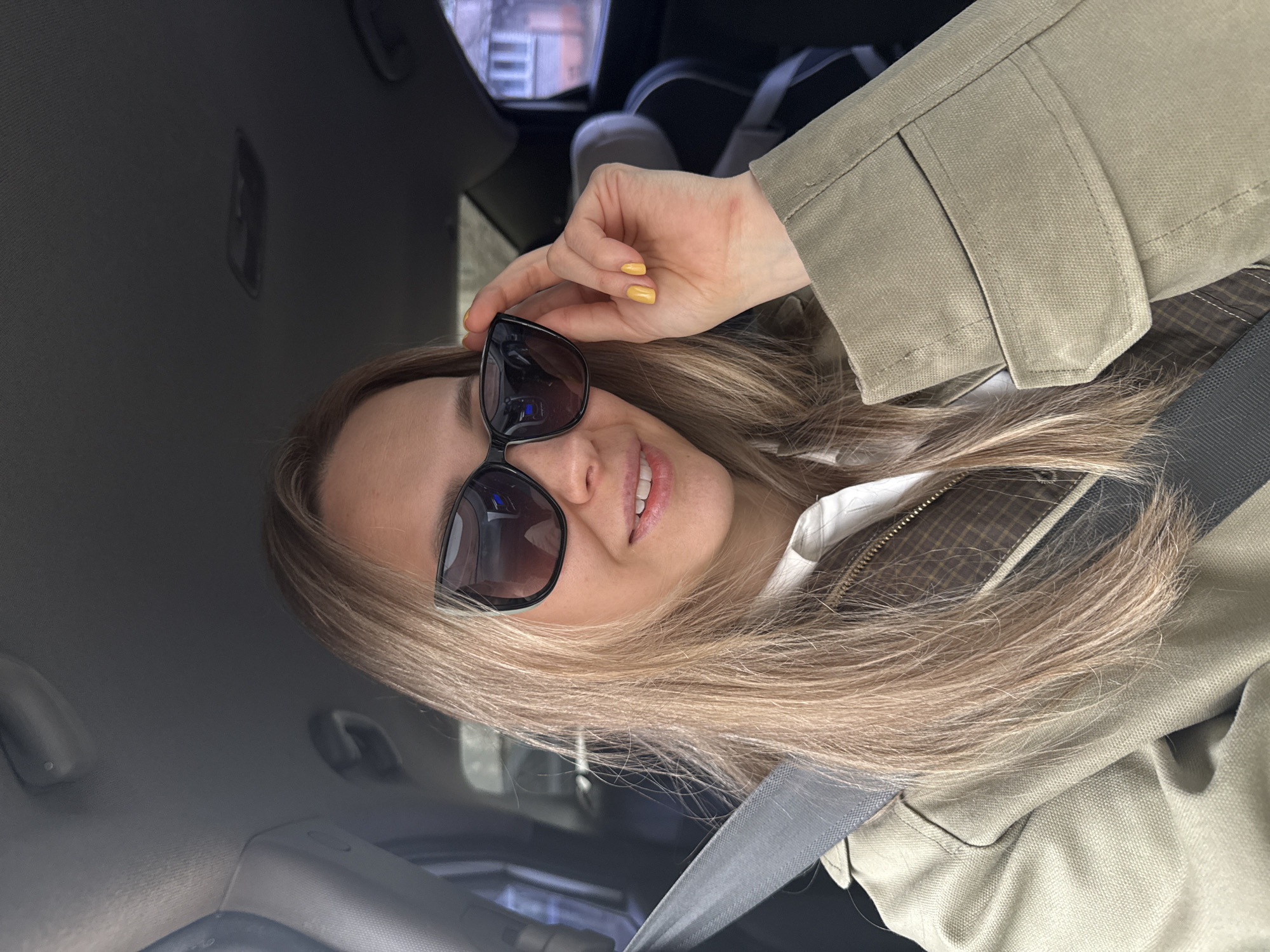 A woman with blonde hair is seated in a car, wearing sunglasses and a tan jacket. She is holding the sunglasses with her left hand, which has yellow nail polish. She is wearing a seatbelt. The location is inside the car with an out of focus background, so the exact city in Chaikovsky, Russia cannot be determined.