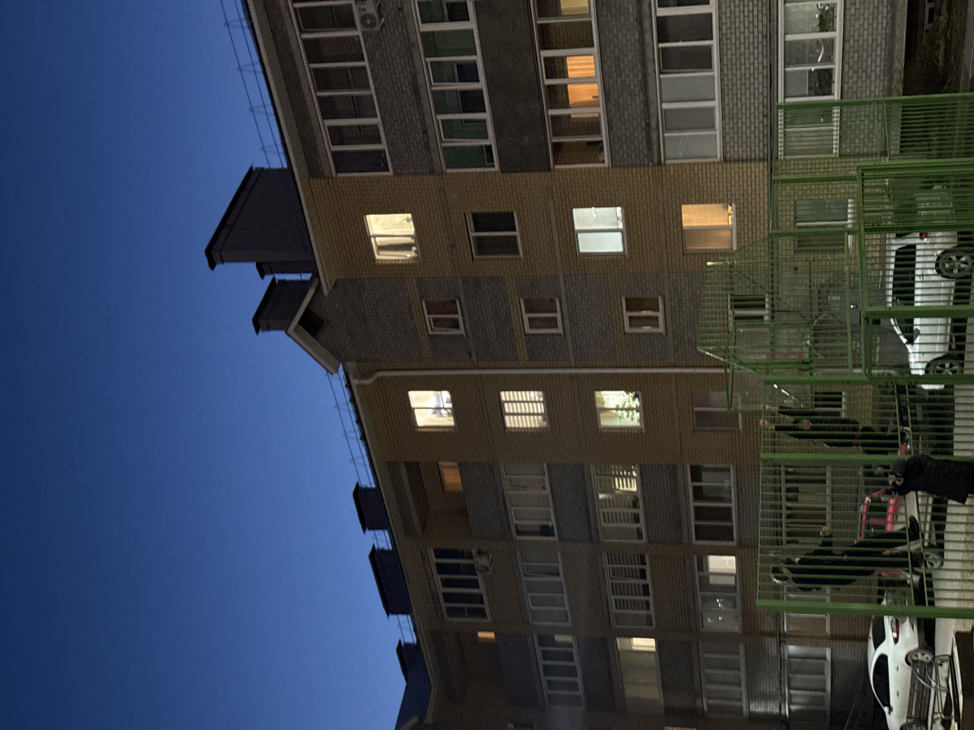 The image shows a multi-story brick building in Votkinsk, Russia, at dusk. Several windows are illuminated, indicating activity inside. A green metal fence partially obscures the view of the building's right side, where cars are parked. People are visible within the confines of the fence. The sky is a deep blue.
