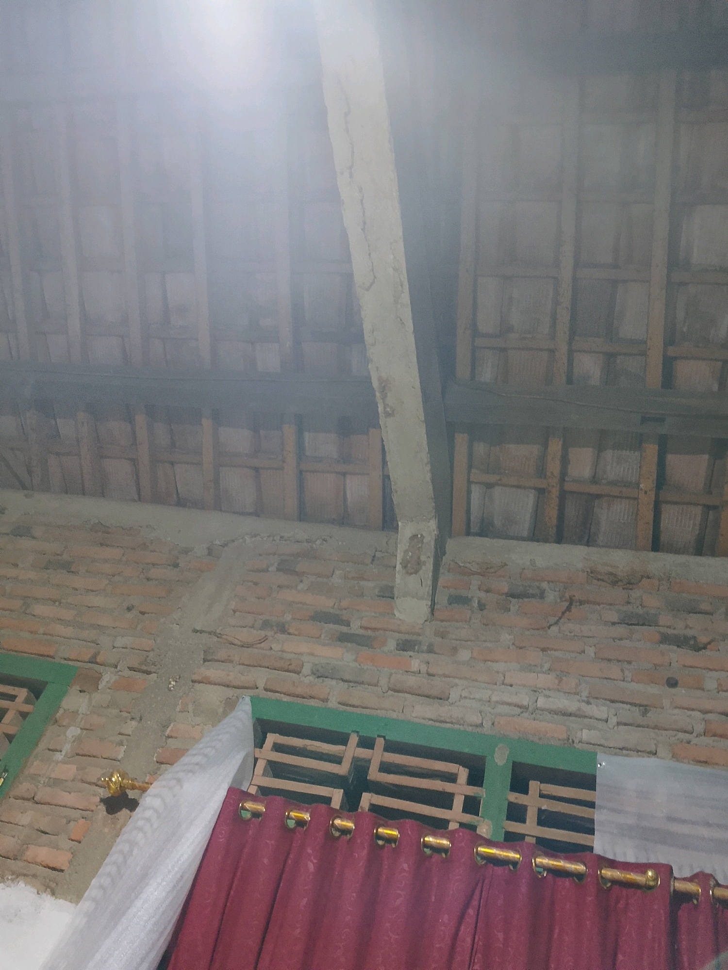 The image displays the interior of a building under construction or renovation in Negeri Agung, Indonesia. The ceiling structure is partially visible, with wooden beams and what appears to be tile roofing. A prominent concrete beam runs diagonally across the upper portion of the image. The walls are constructed of brick. A green-framed window is visible, with wooden slats behind it. Partially covering the window area are maroon curtains with gold grommets and a white sheer curtain. A gold decorative element, possibly a curtain tie-back or part of a fixture, is attached to the wall near the white curtain. The lighting in the image is somewhat hazy, suggesting either dust or a bright light source directly above and to the left of the frame. The overall scene depicts a functional, unfinished interior space.